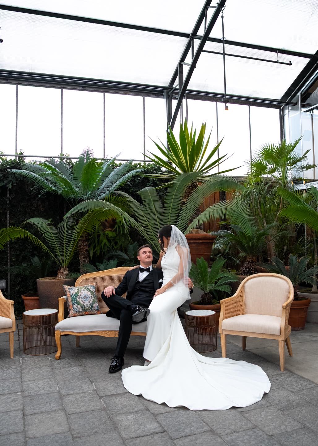 Bride and groom sitting on a sofa in a greenhouse with large potted plants and chairs, bride in white gown, groom in black suit.