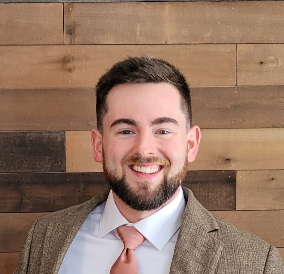 Portrait of a smiling man with dark hair and beard, wearing a suit and tie, standing in front of a wooden wall.