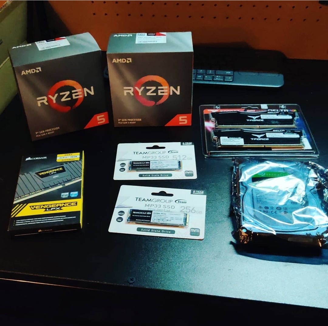 Computer components and accessories on a black desk, including AMD Ryzen 5 boxes, RAM sticks, SSDs, a cooling fan, and a keyboard.