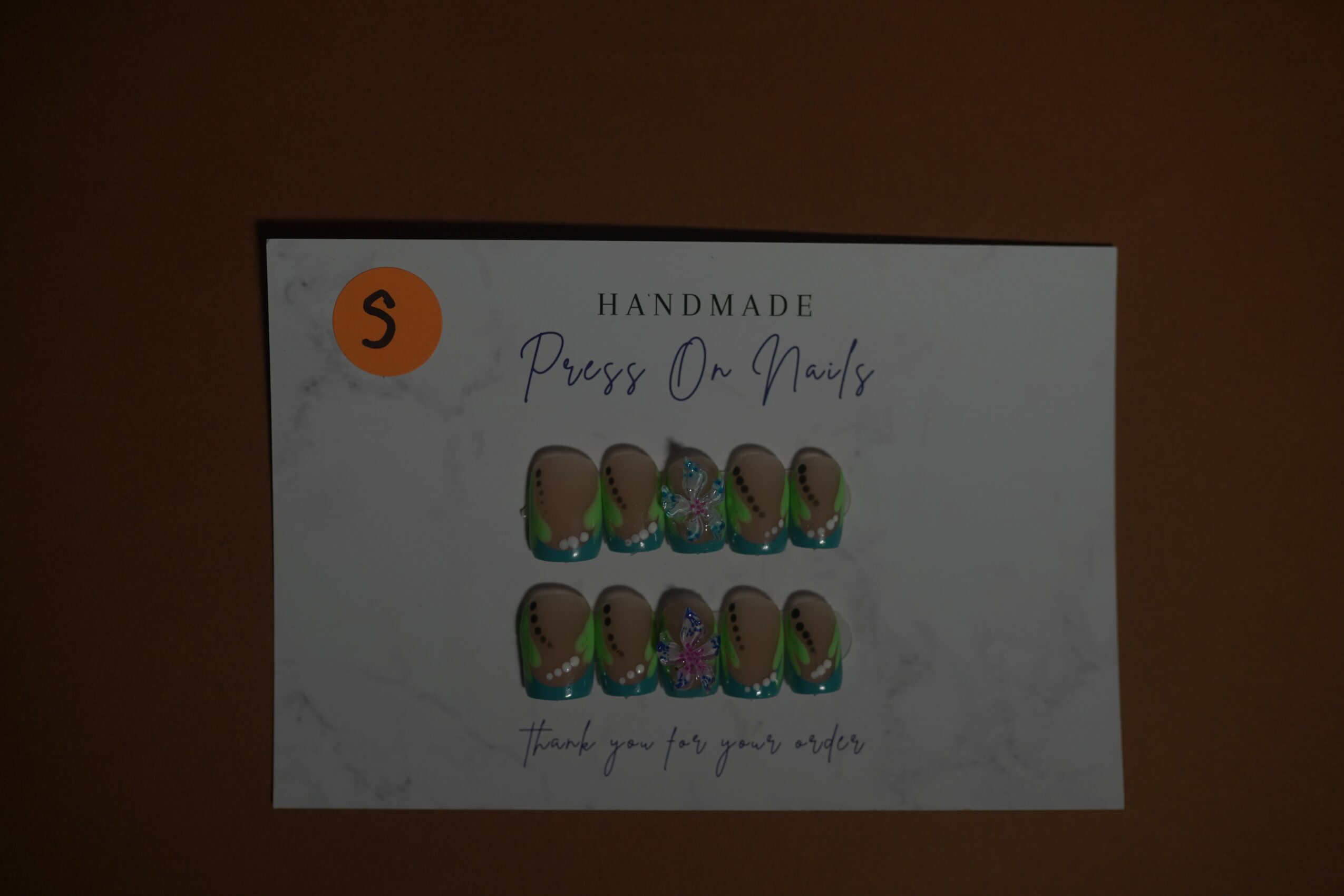 Business card with handmade nail art images, orange circle with 'S', and text about press-on nails, on a dark background.