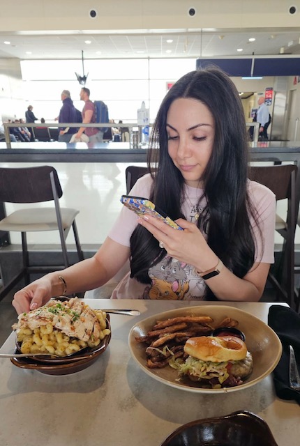 Young woman with long dark hair looking at her phone while sitting at a table with two plates of food, in a bright indoor setting.