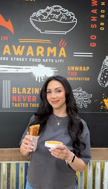 Young woman holding a drink and a snack in front of a black wall with food-themed illustrations and text.
