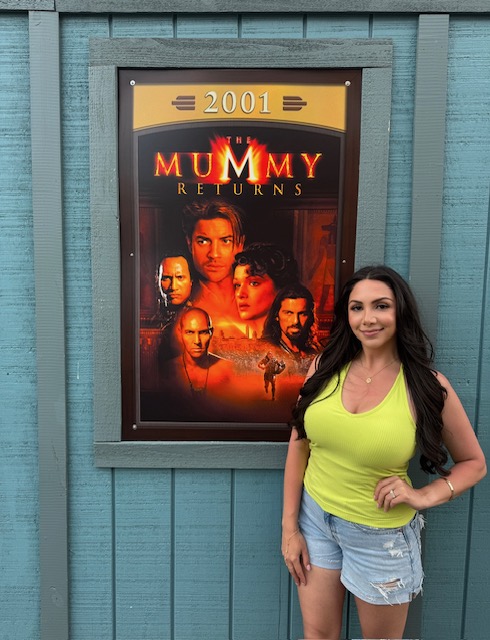 Woman with long dark hair in a yellow tank top and denim shorts standing outside next to a movie poster for 'The Mummy Returns' in a blue wooden frame.