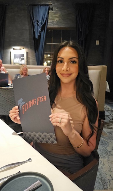 Woman with long dark hair holding a sign that says 'Flying Fish' at a restaurant table.