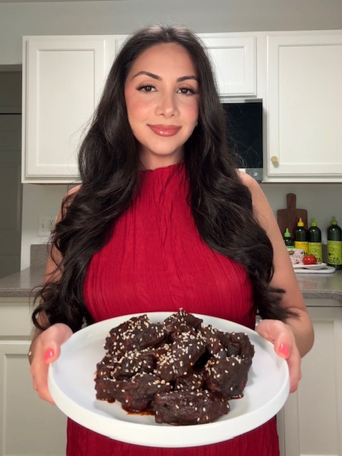Young woman with long dark hair in a red sleeveless top holding a plate of chocolate brownies, smiling in a kitchen.