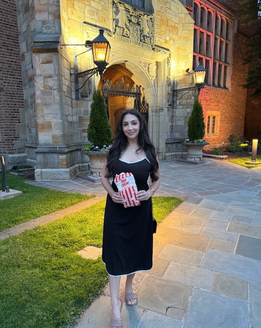 Woman in black dress holding a gift box, standing outside a historic stone building with arched entrance and lanterns.