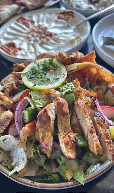 Grilled chicken salad with lemon slice, lettuce, onions, and tomatoes, served with a side of creamy dip or sauce.