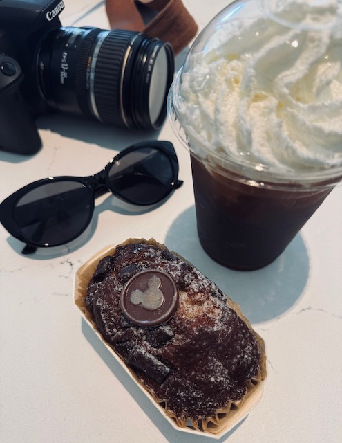A chocolate pastry with a Mickey Mouse chocolate piece on top, a cold drink with whipped cream, sunglasses, and a camera on a white surface.
