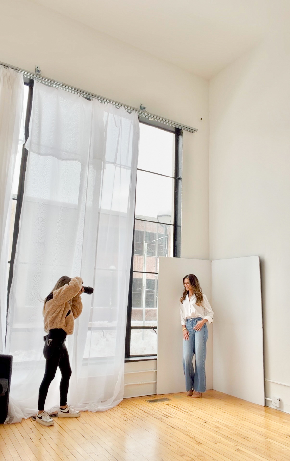 Photographer taking picture of woman standing near window with white curtains, sunlight streaming in, wooden floor, plain white wall.