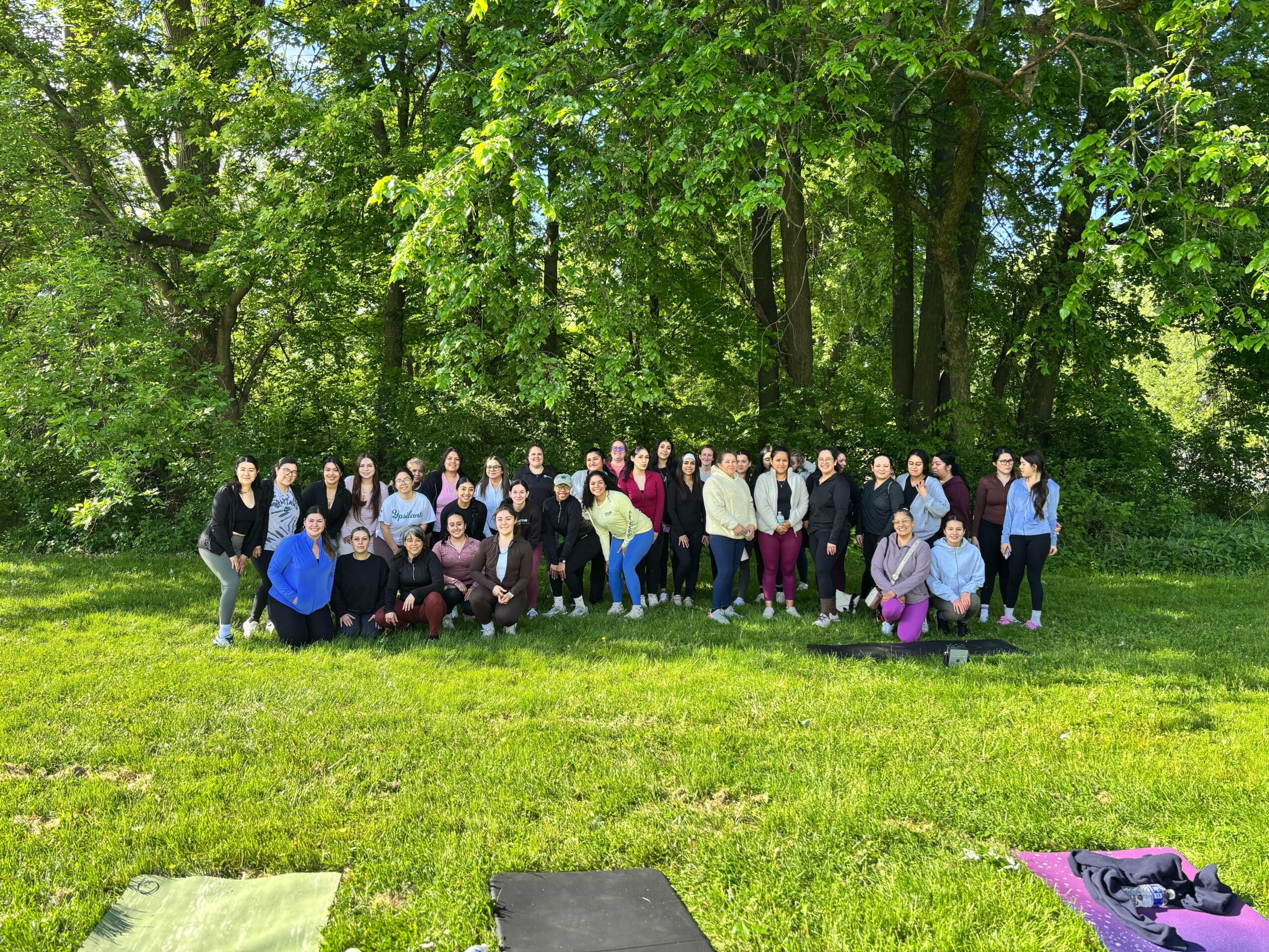 Group of people standing outdoors on grass with trees in the background, sunny day.
