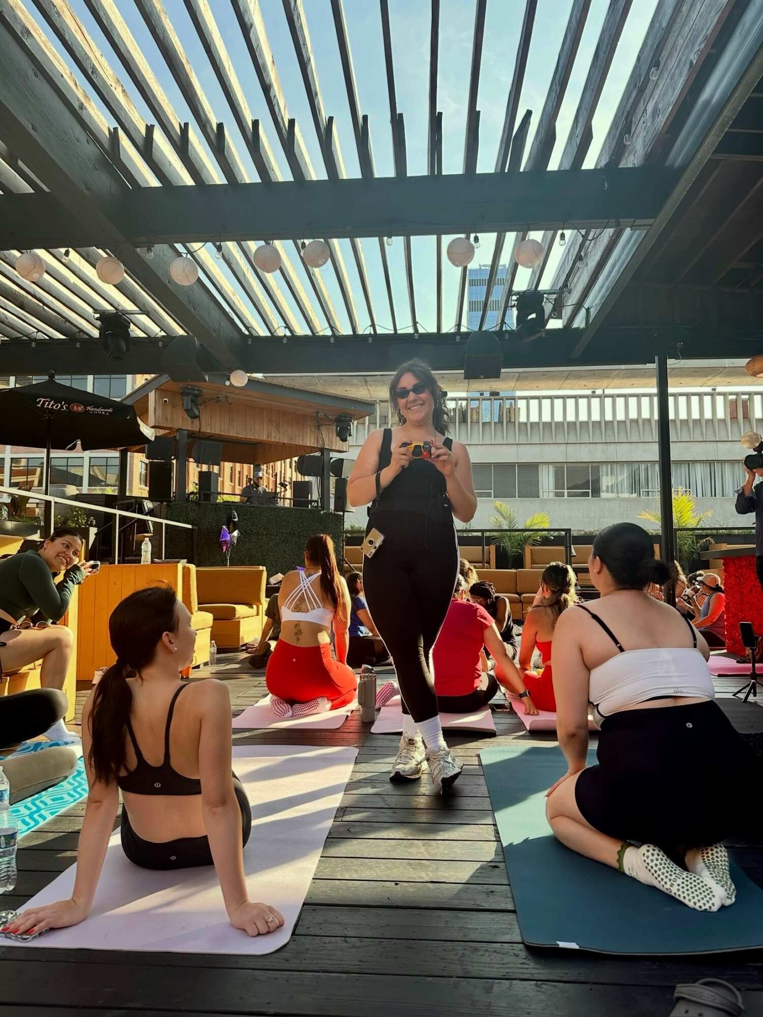 Woman standing on a platform taking a photo, surrounded by seated people on mats outdoors, with a modern building and sky overhead.