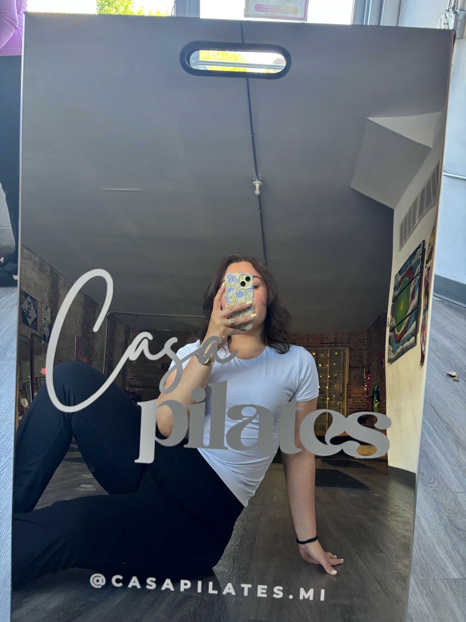 Person taking a mirror selfie with a phone, sitting on the floor, wearing a white top and black pants.
