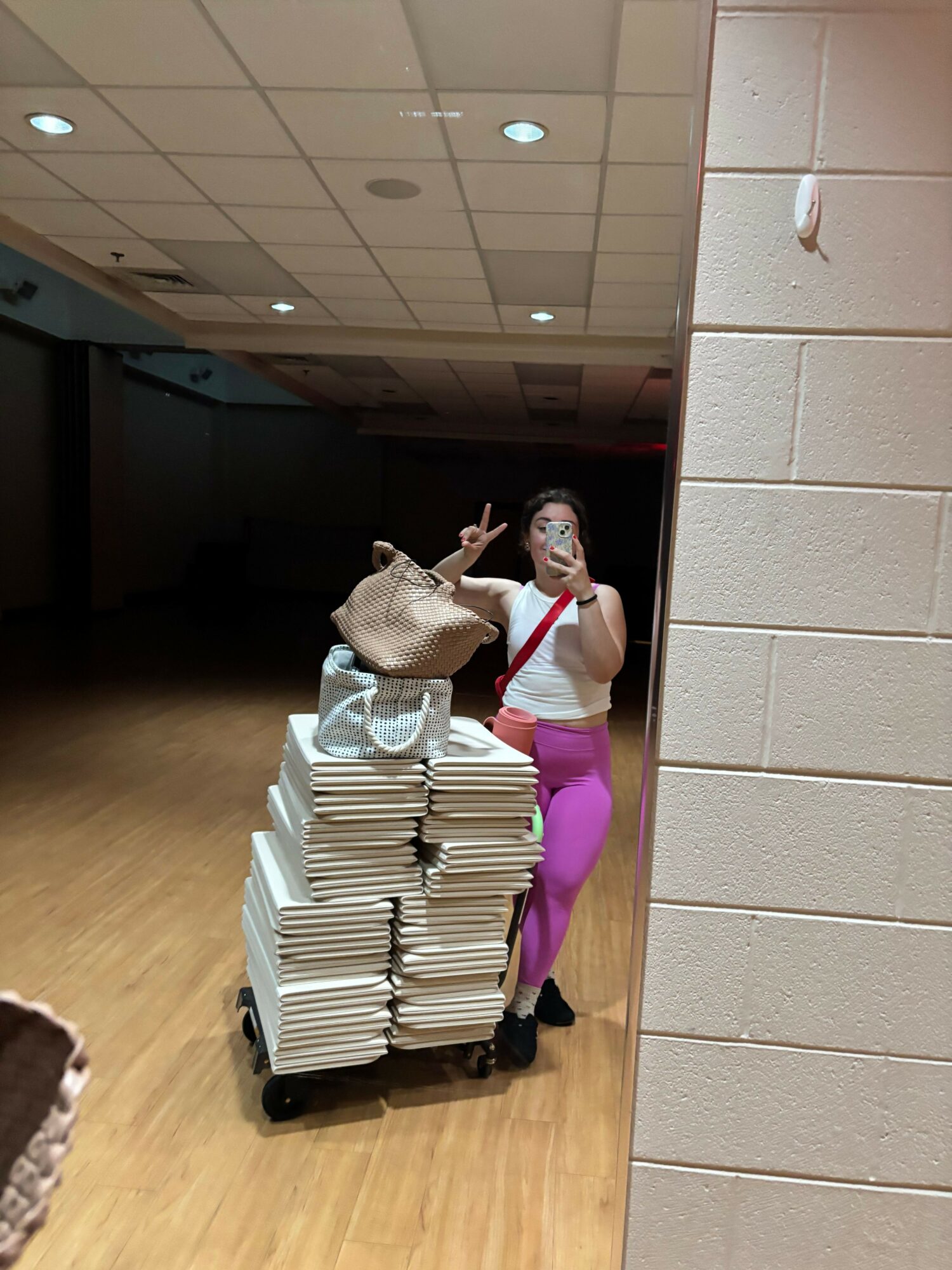 Two women stand next to a stack of books on a wooden floor, one making a peace sign, the other holding a bag.