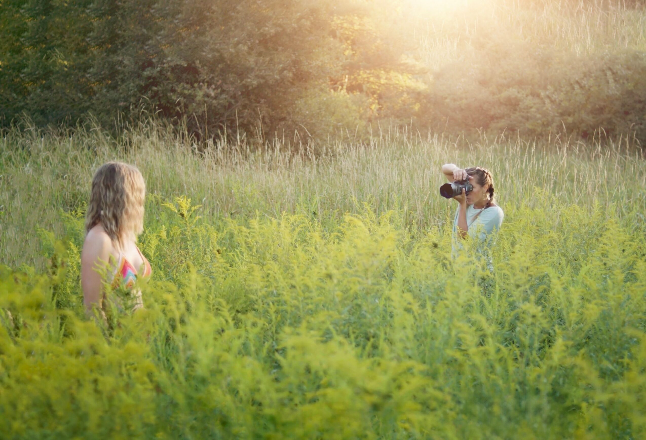 Two women in a grassy field, one taking a photo of the other, with sunlight shining from the top right.