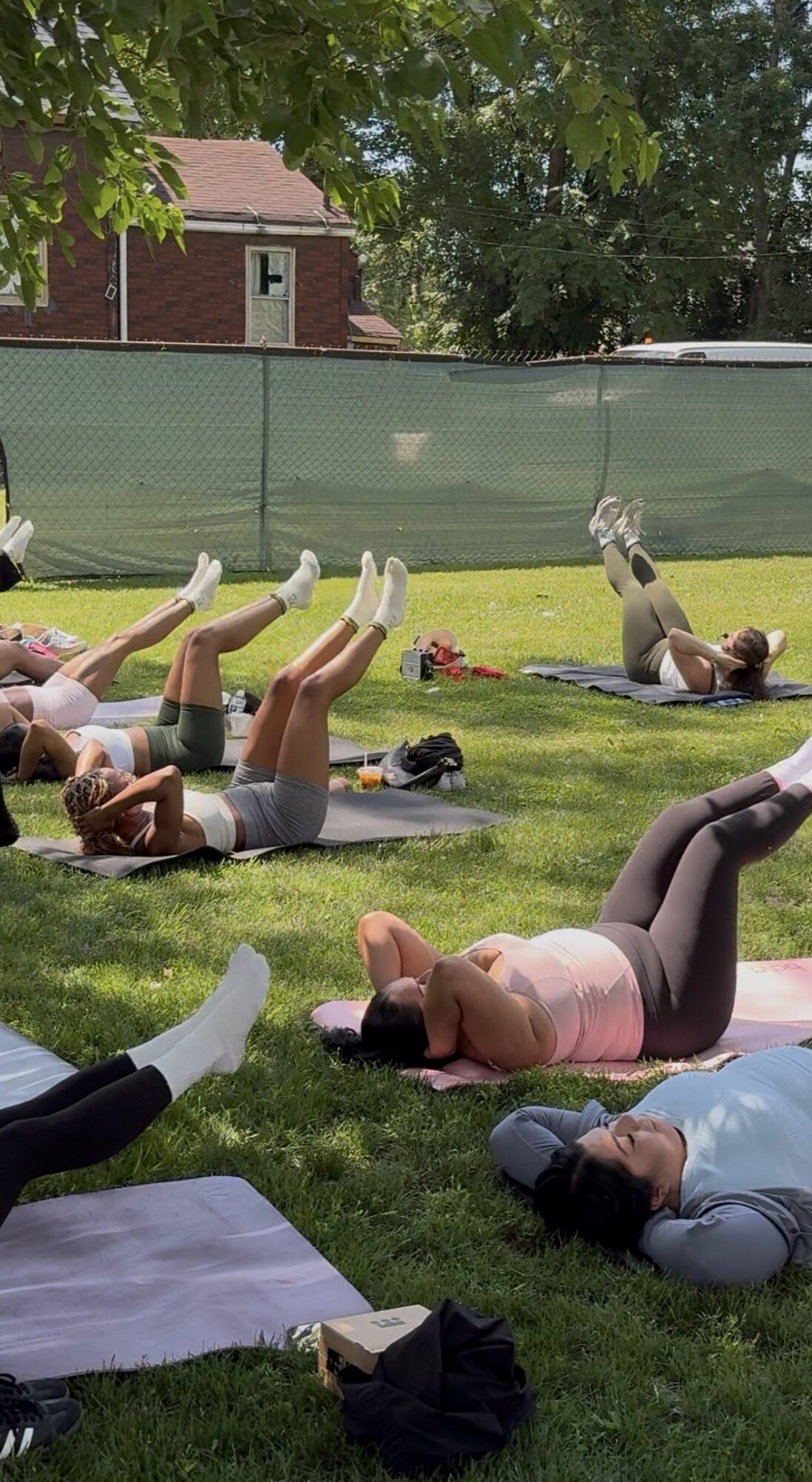 People lying on mats outdoors, participating in a group exercise or yoga session on grass, with trees and a building in background.