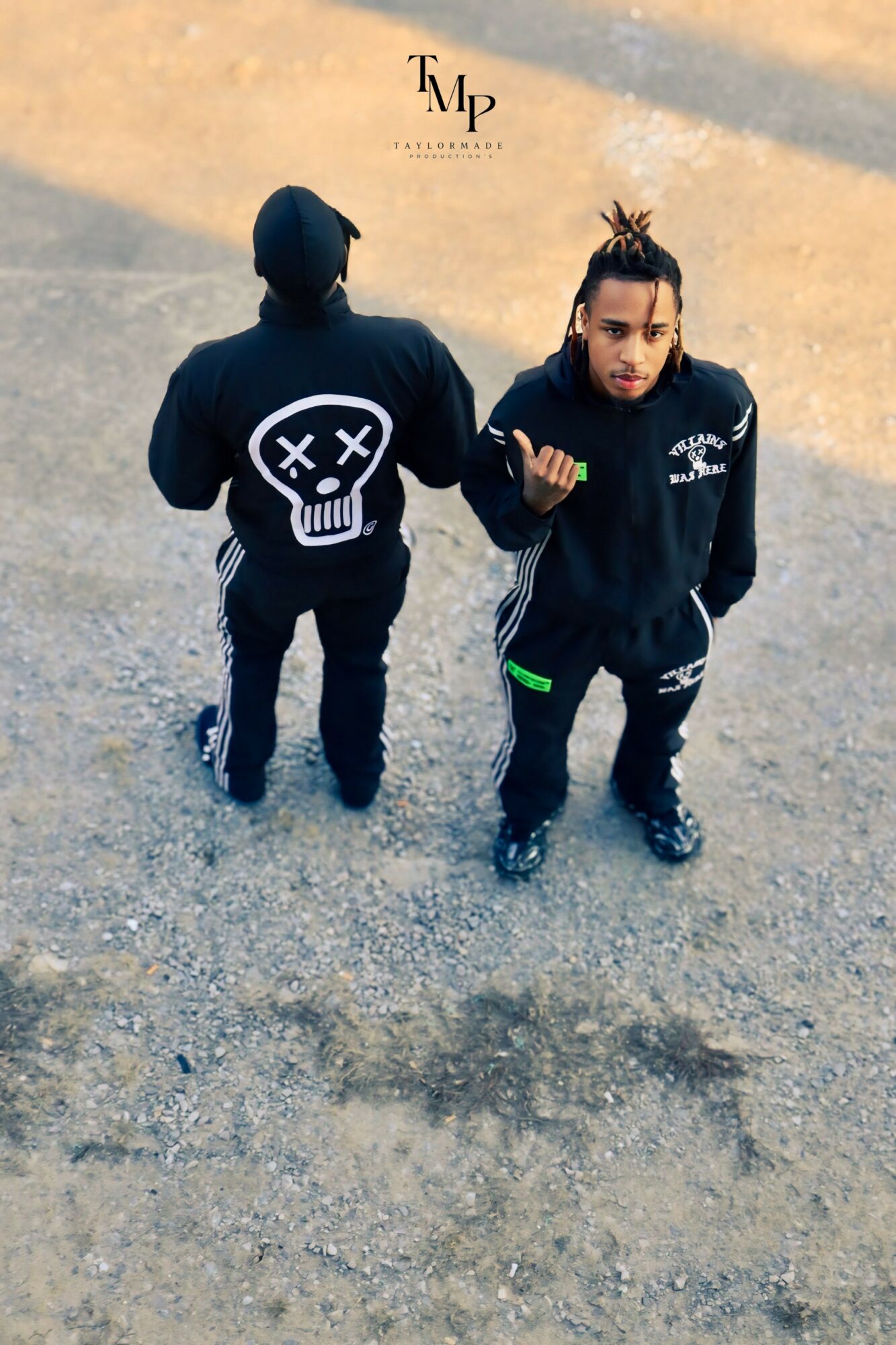 Two young men stand on a street, one facing away with a hoodie featuring a skull design, the other facing forward with a hand gesture.