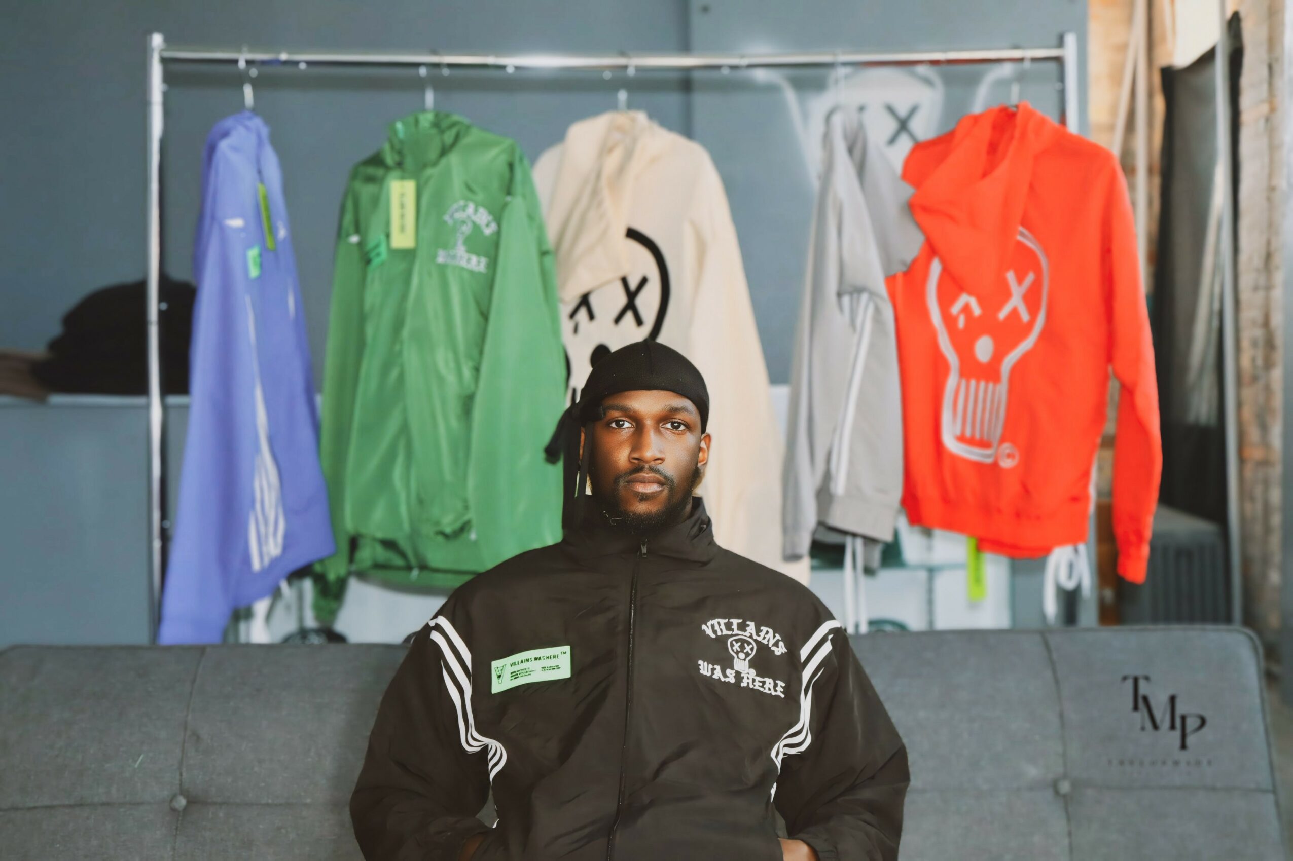 Person with black headwear and black jacket sitting in front of clothing display with colorful hoodies and sweatshirts, some with graphic designs.