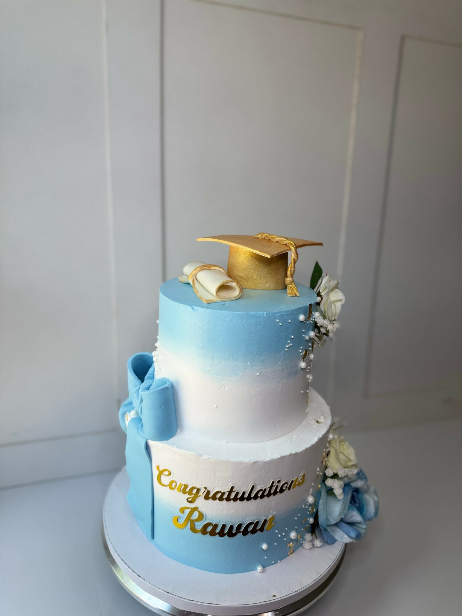 Two-tiered cake with blue and white gradient, decorated with a gold graduation cap, scroll, and blue bows, on a white base.