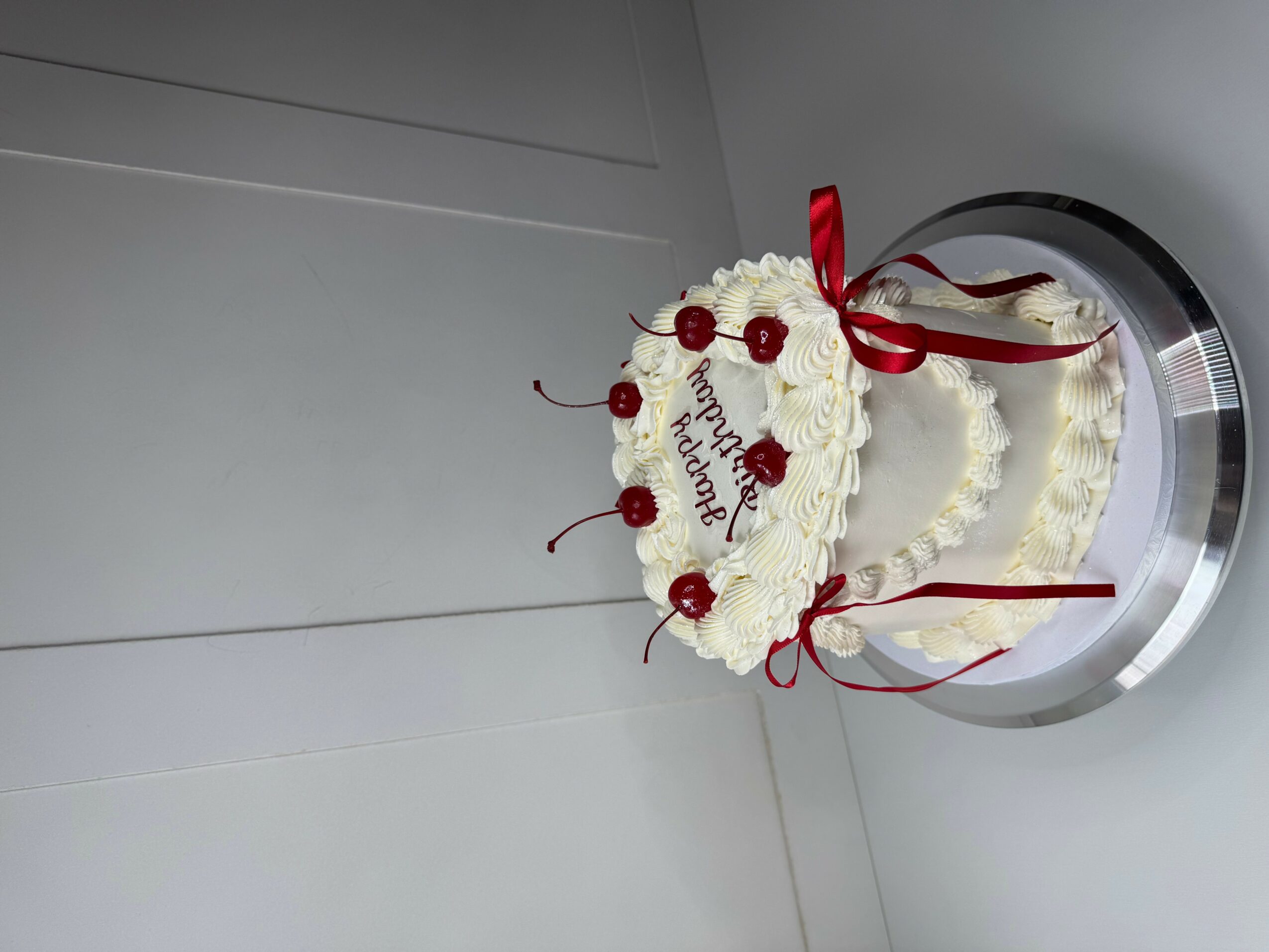 A white cake decorated with red ribbons and cherries, with a message on top, on a silver stand.