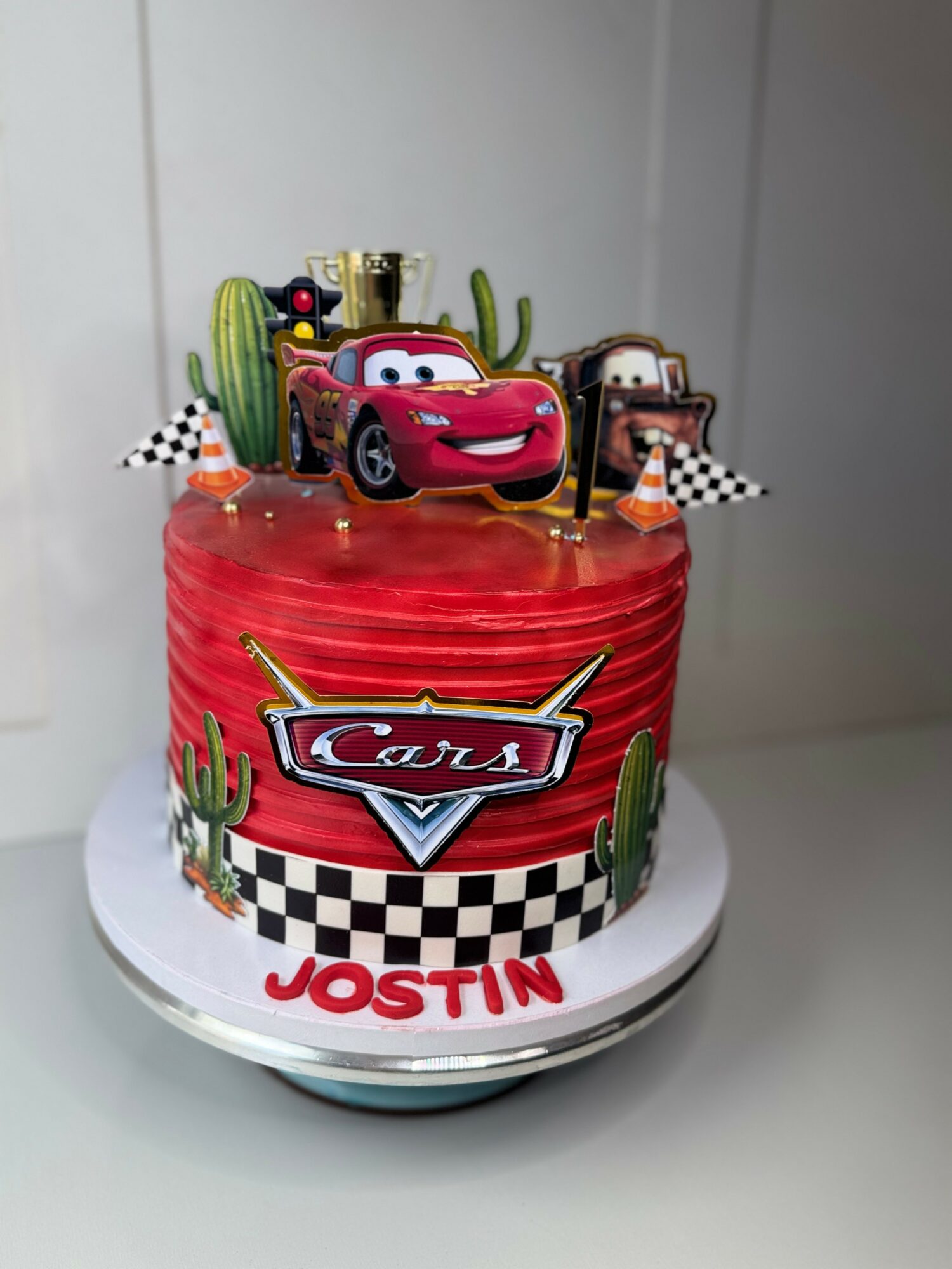 Cake decorated with Cars characters, cacti, checkered flags, and a name Jostin, with a red and black theme.