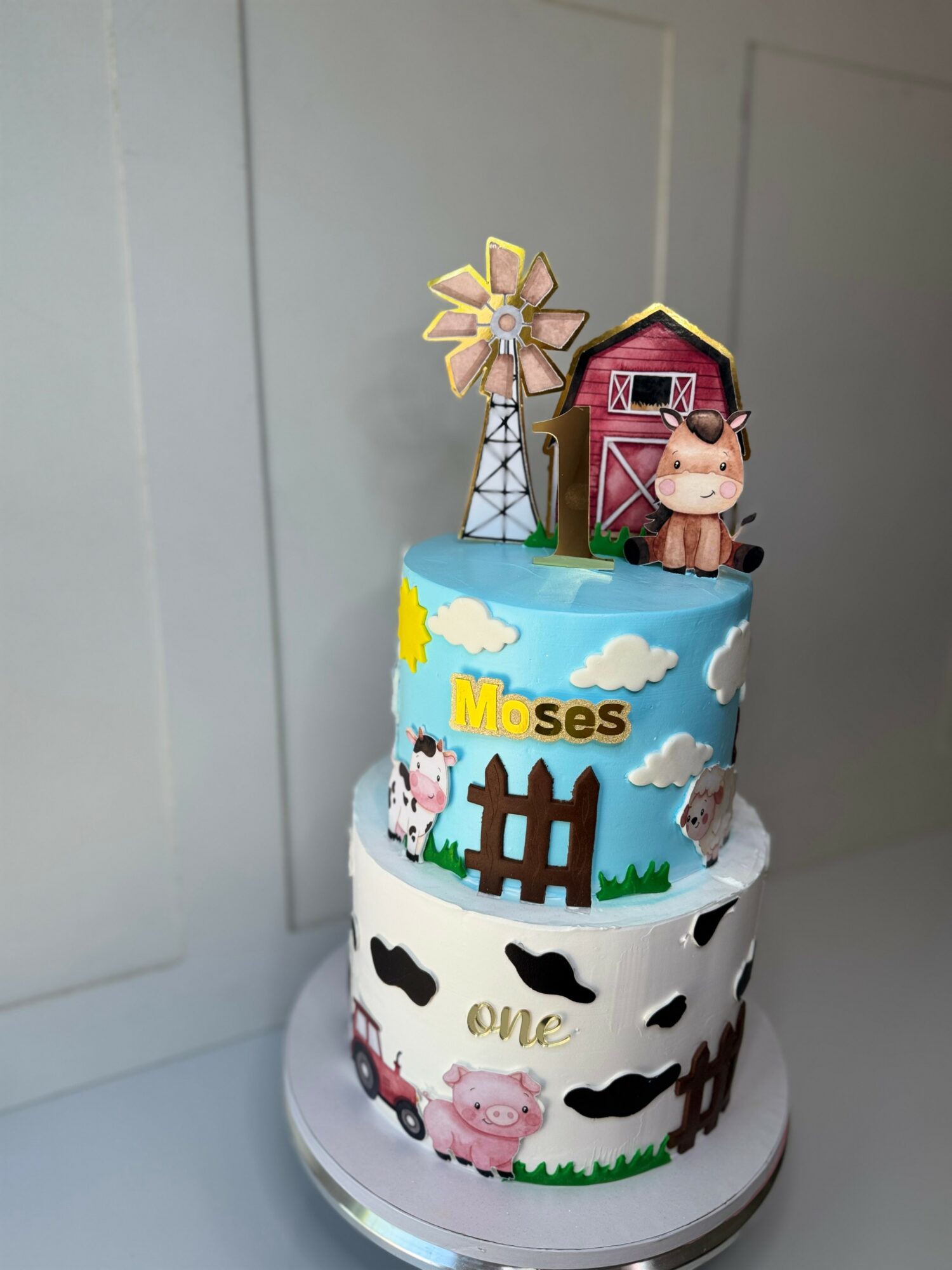 Two-tiered cake with farm animal decorations, a windmill, and a barn topper, featuring clouds, animals, and the name Moses.