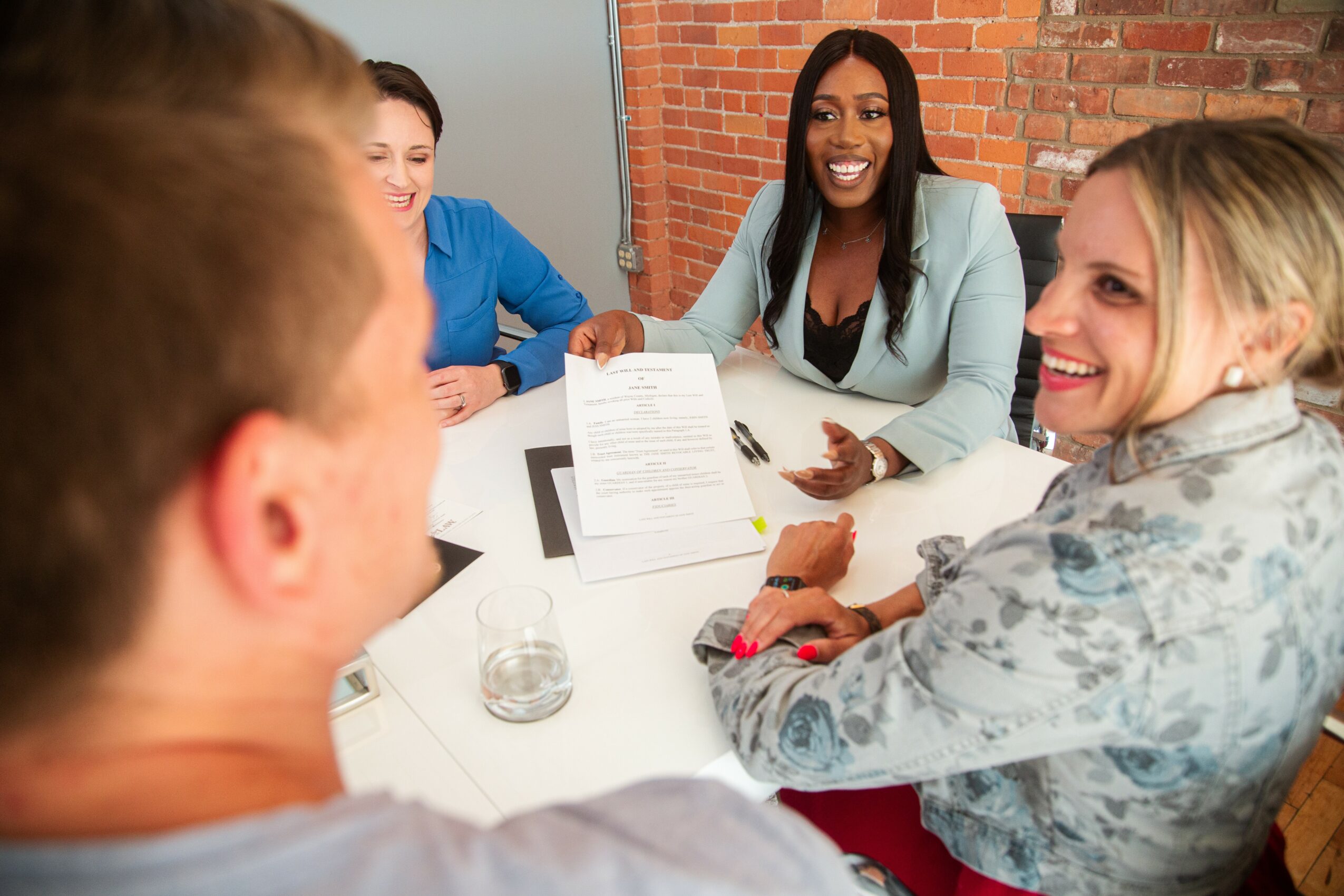 Group of five people sitting around a table, smiling and talking, with documents and a glass of water on the table.
