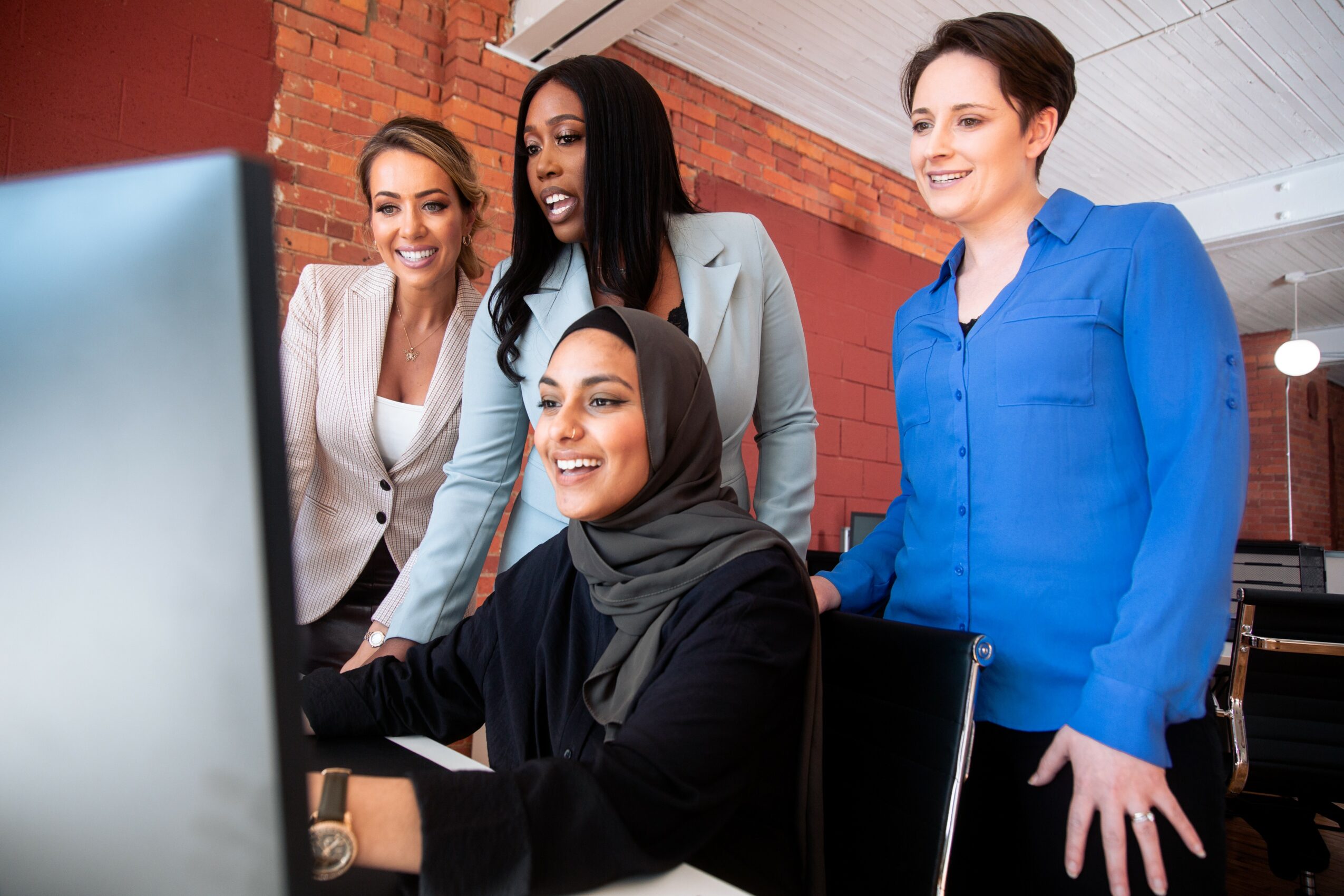 Four women gathered around a computer, smiling and looking at the screen in a modern office setting.