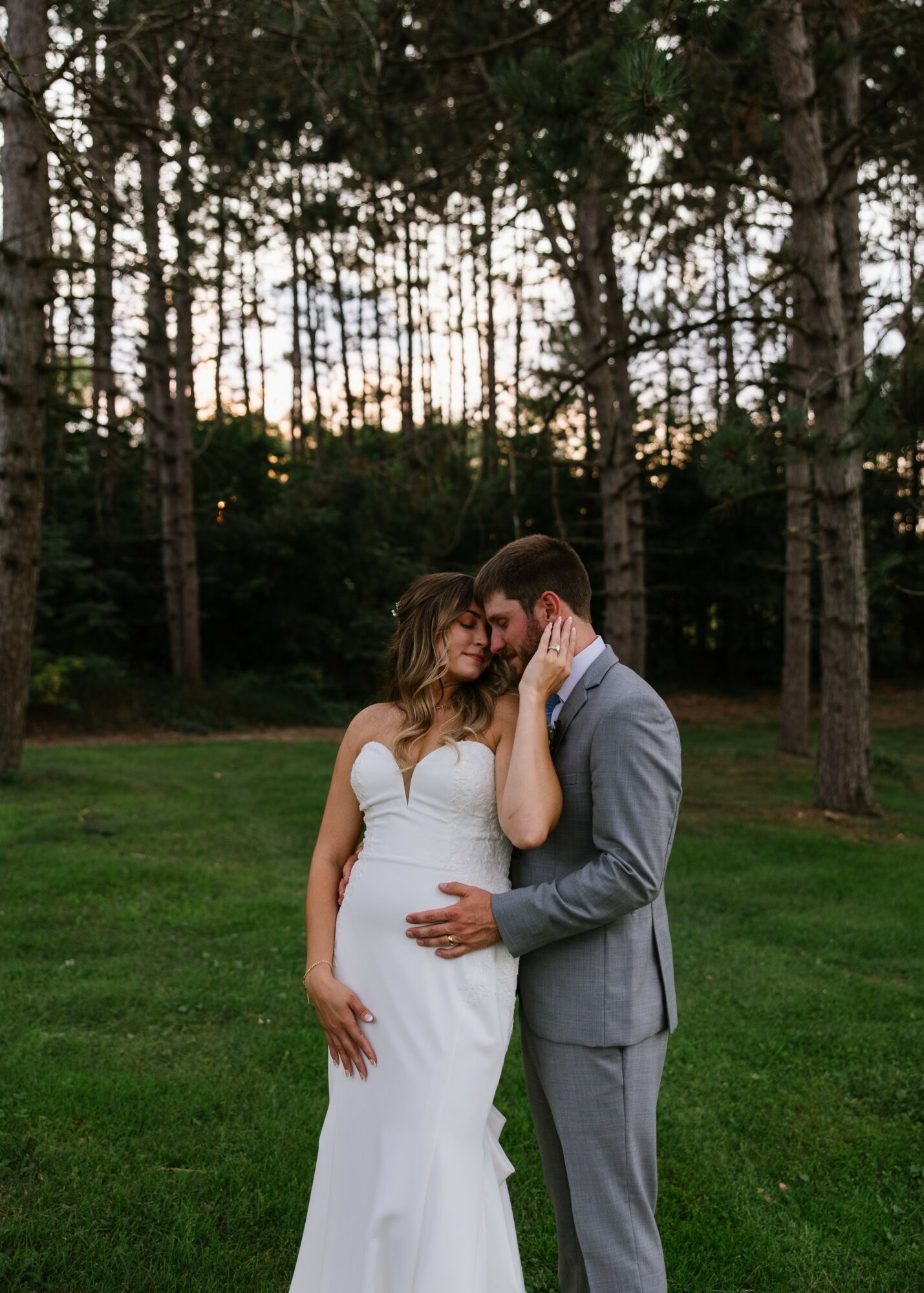 A couple stands close together on a grassy area with trees in the background, embracing each other affectionately.