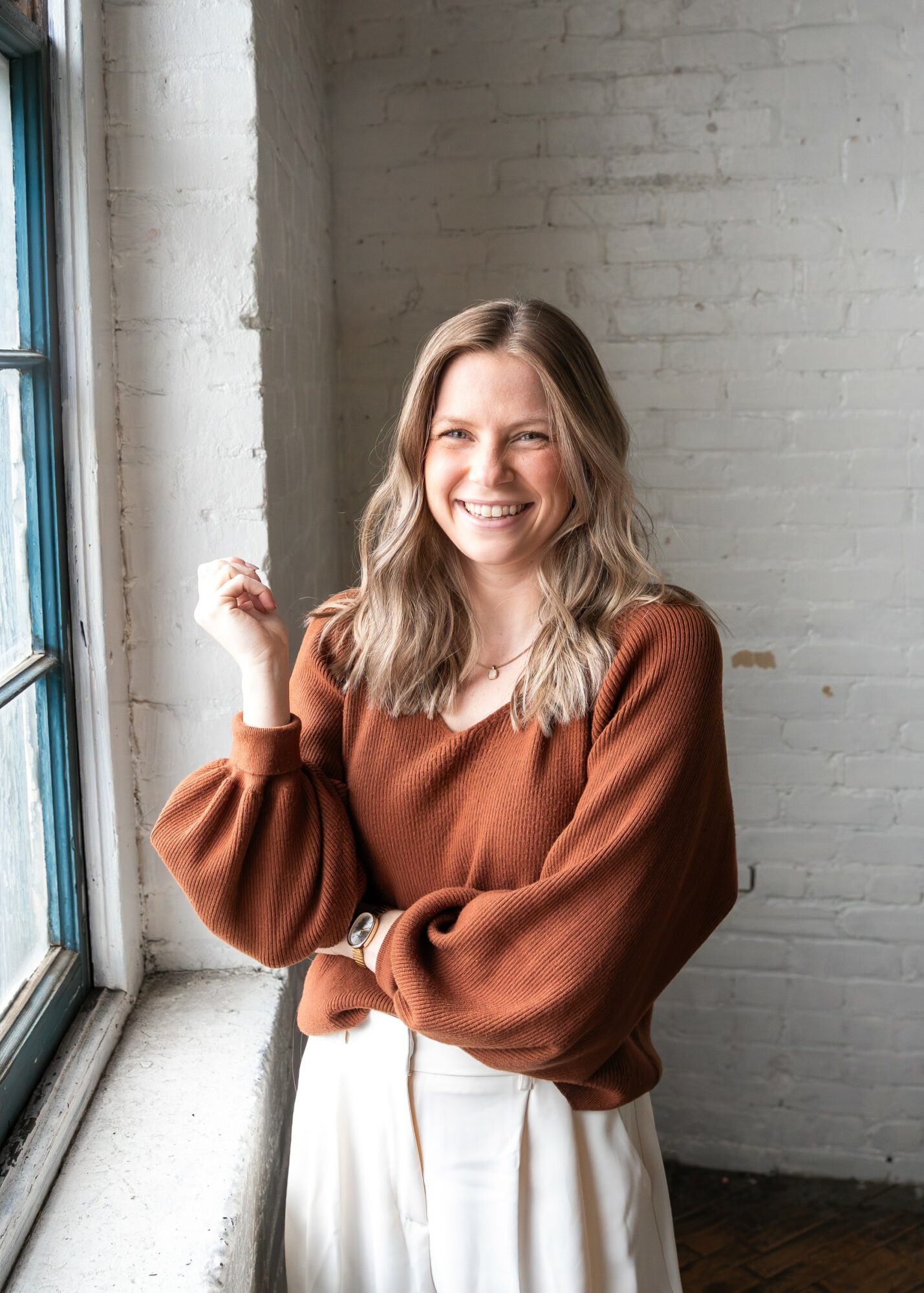 Smiling woman with wavy hair wearing a brown sweater and white pants, standing near a window with a brick wall background.