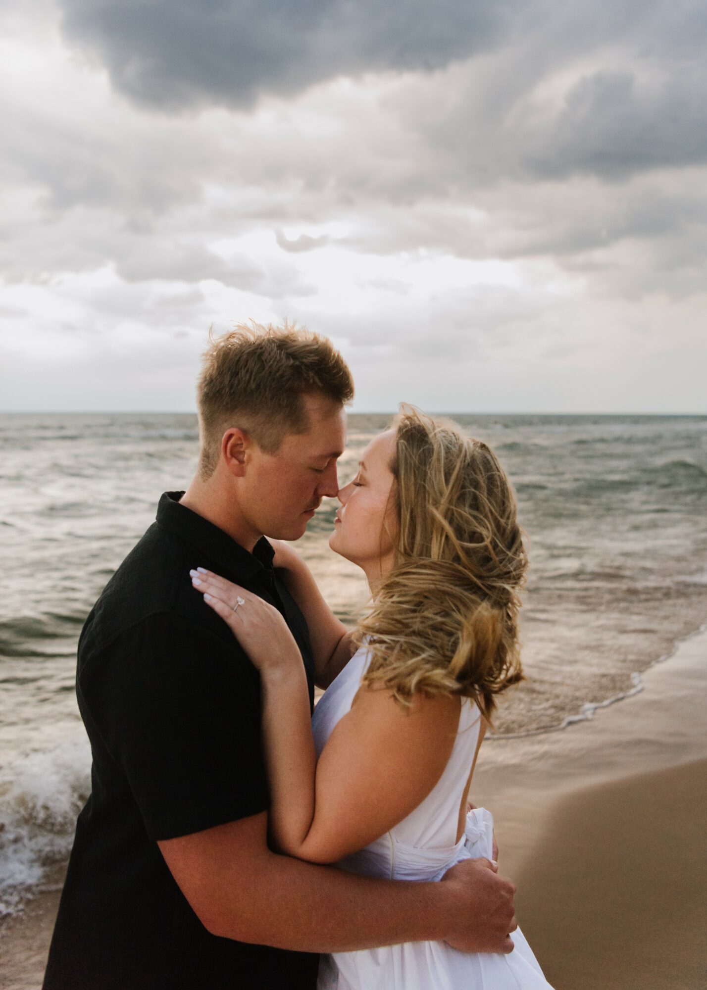 A couple stands close on a beach, facing each other with cloudy sky and ocean in background.
