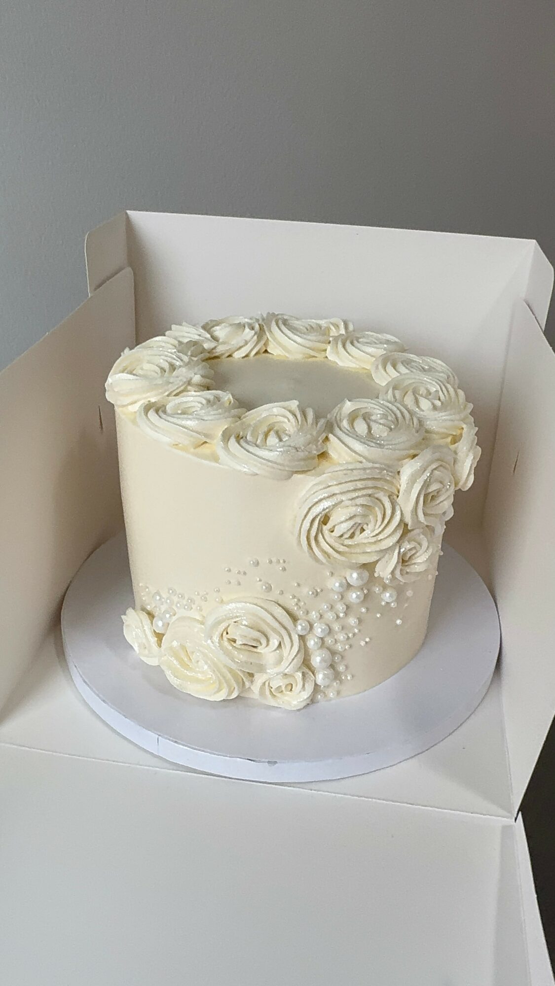 Round white cake decorated with cream roses and small white pearls, inside a white box.