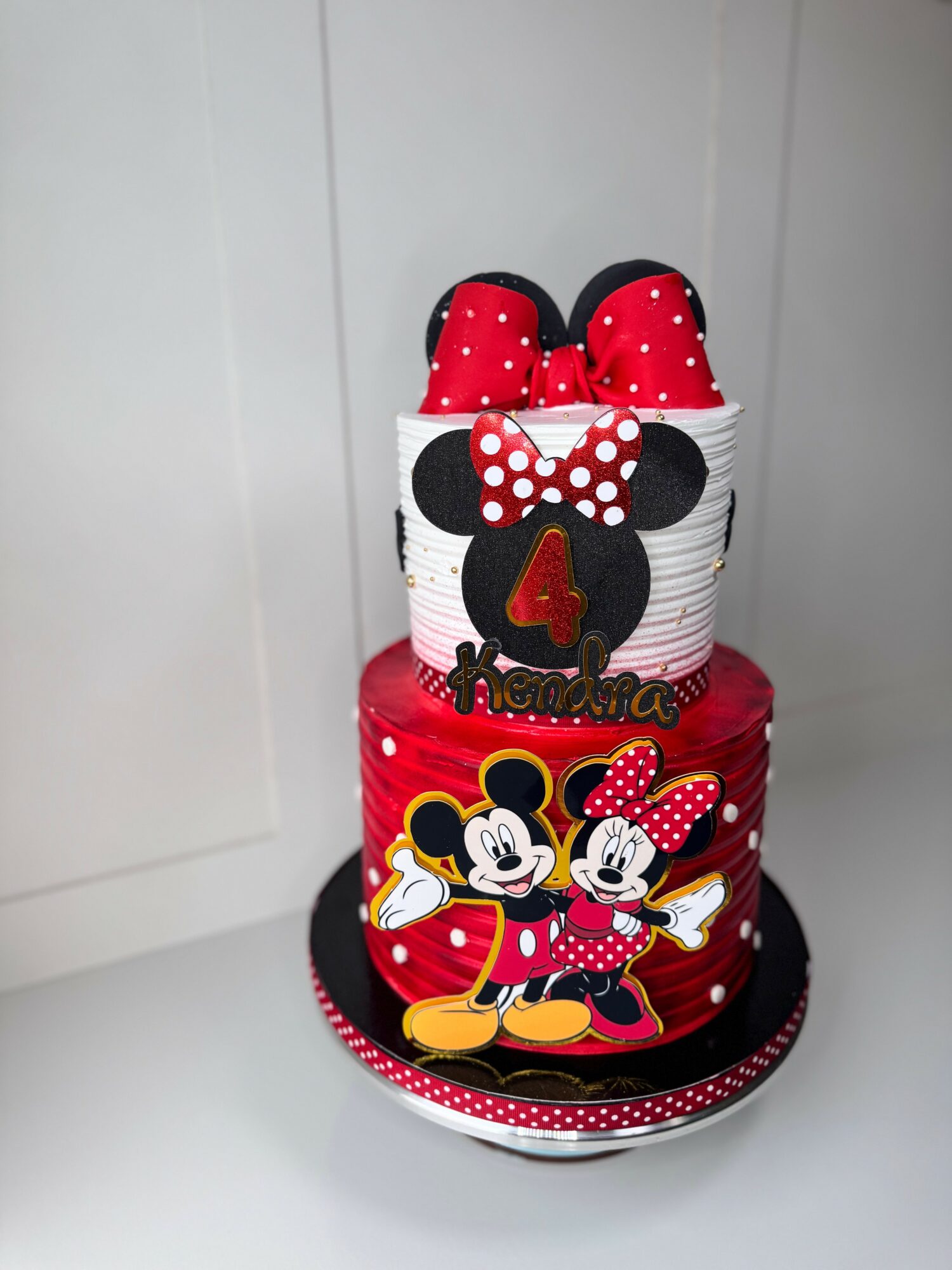 Two-tiered birthday cake with Minnie Mouse theme, featuring Minnie and Mickey illustrations, red, black, and white colors, and a large bow on top.