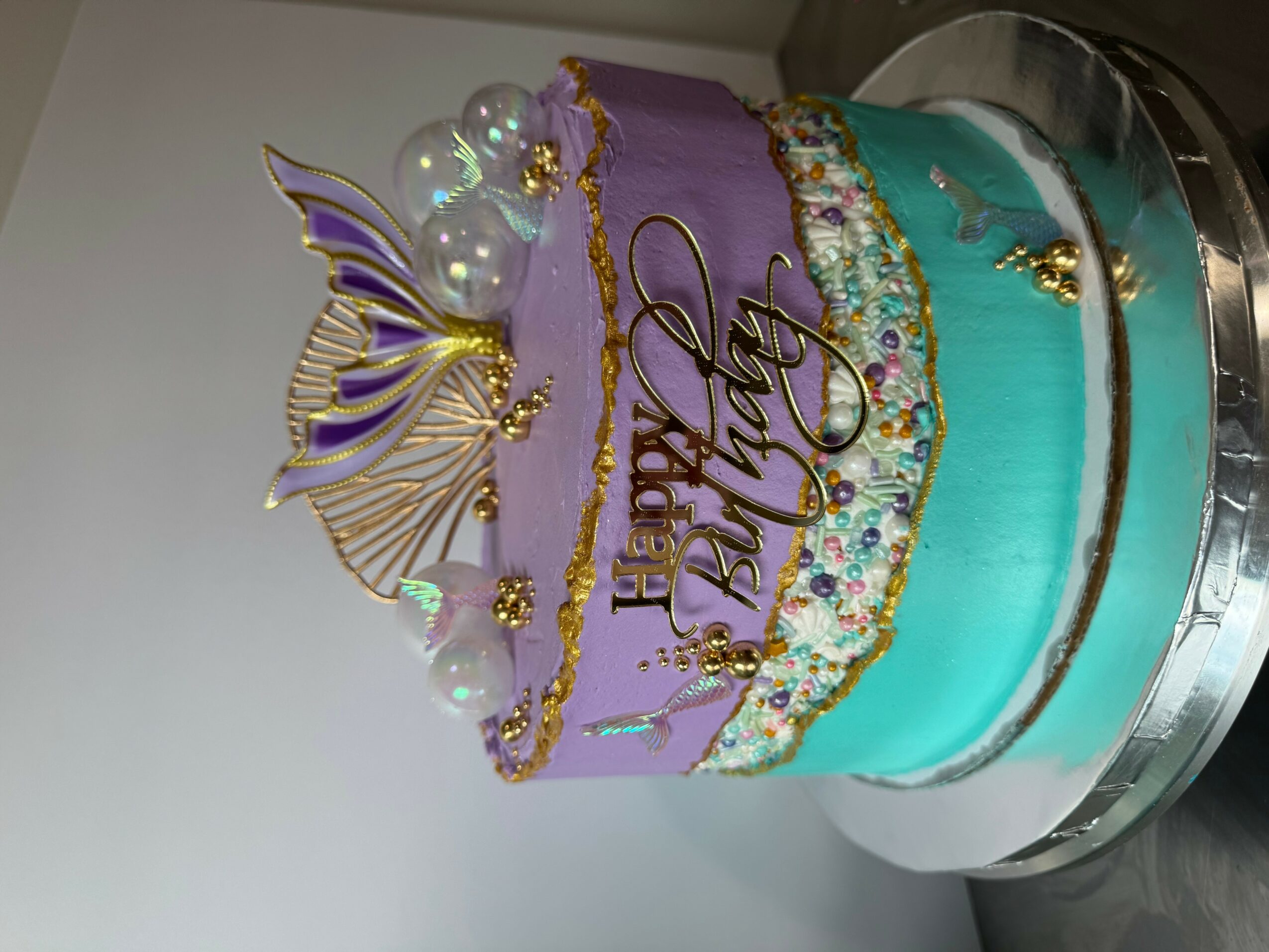 Decorative birthday cake with purple, turquoise, and gold accents, featuring a butterfly topper and pearl decorations.