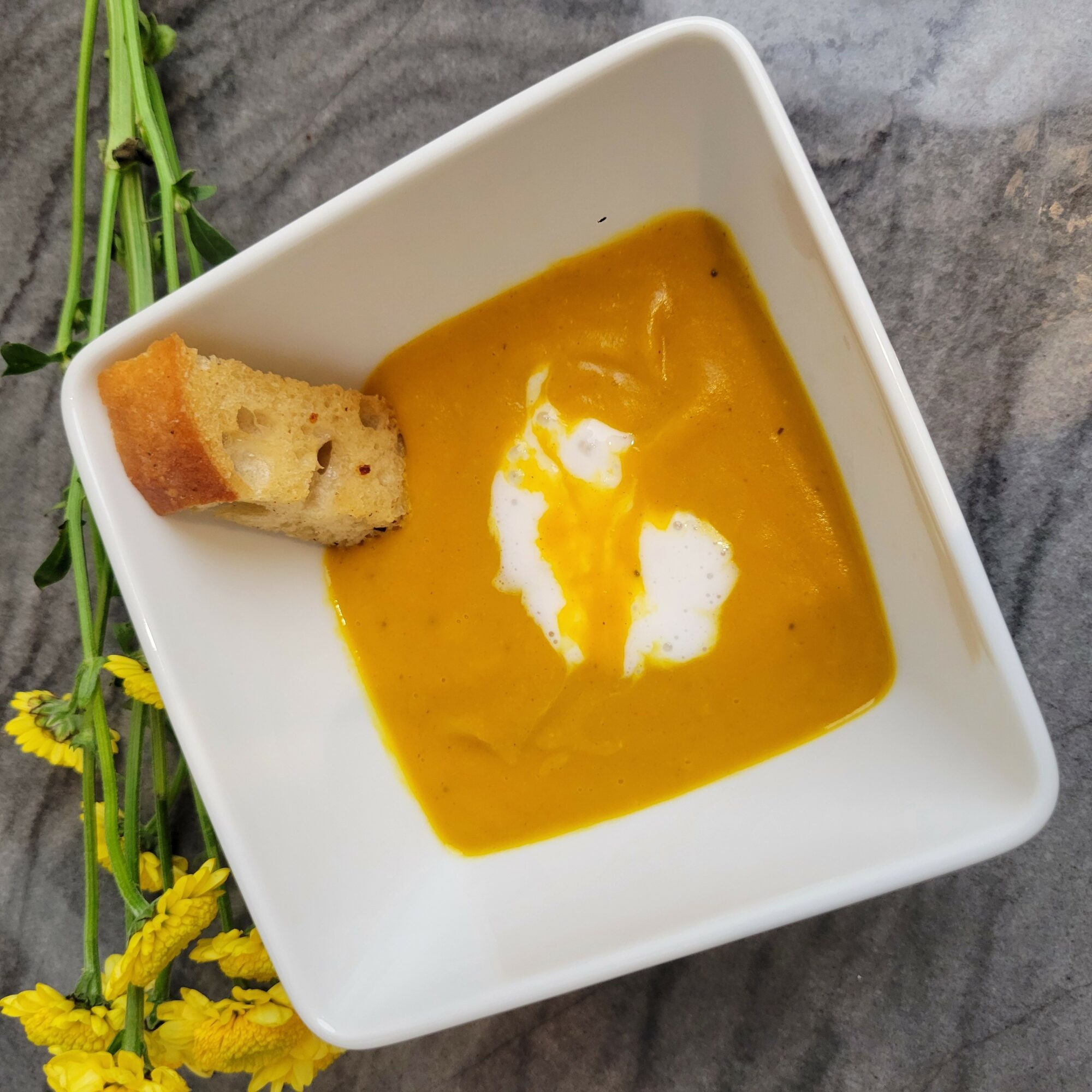 Bowl of orange soup with a dollop of cream and a piece of bread, on a gray surface with yellow flowers nearby.
