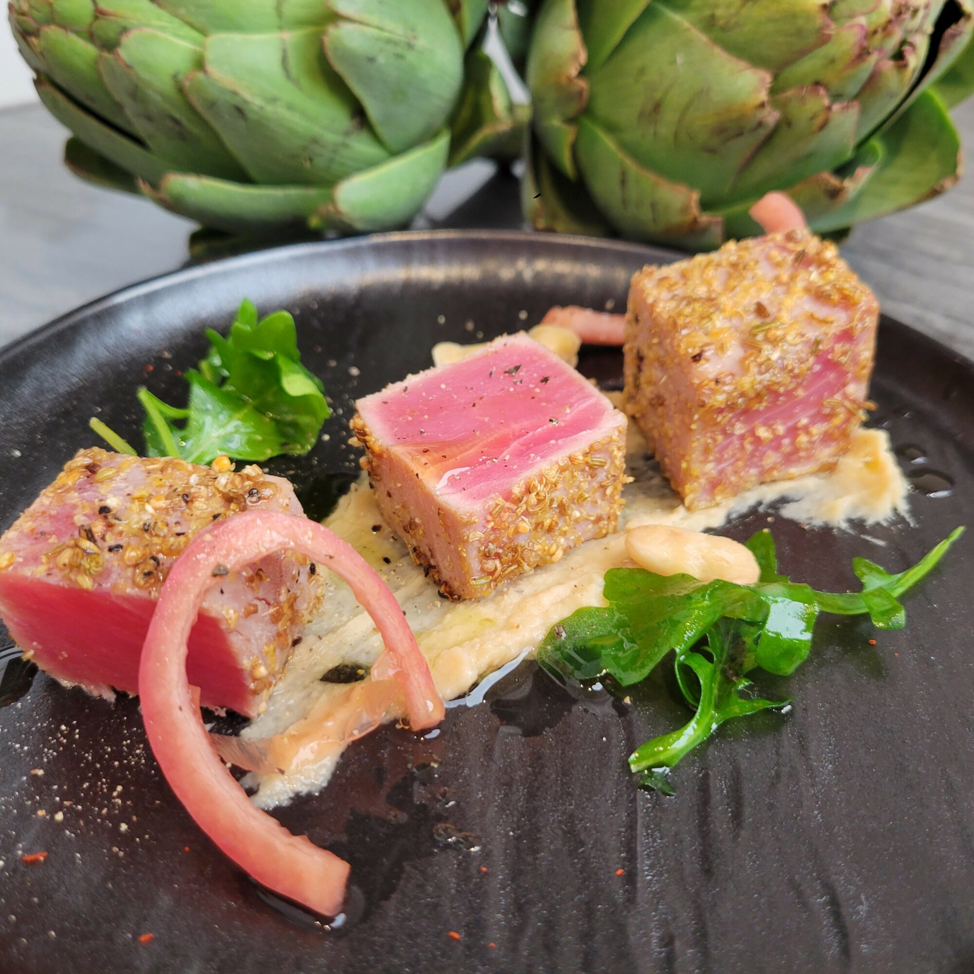 Seared tuna with pink interior, garnished with greens and pickled onions on a black plate, with artichokes in background.