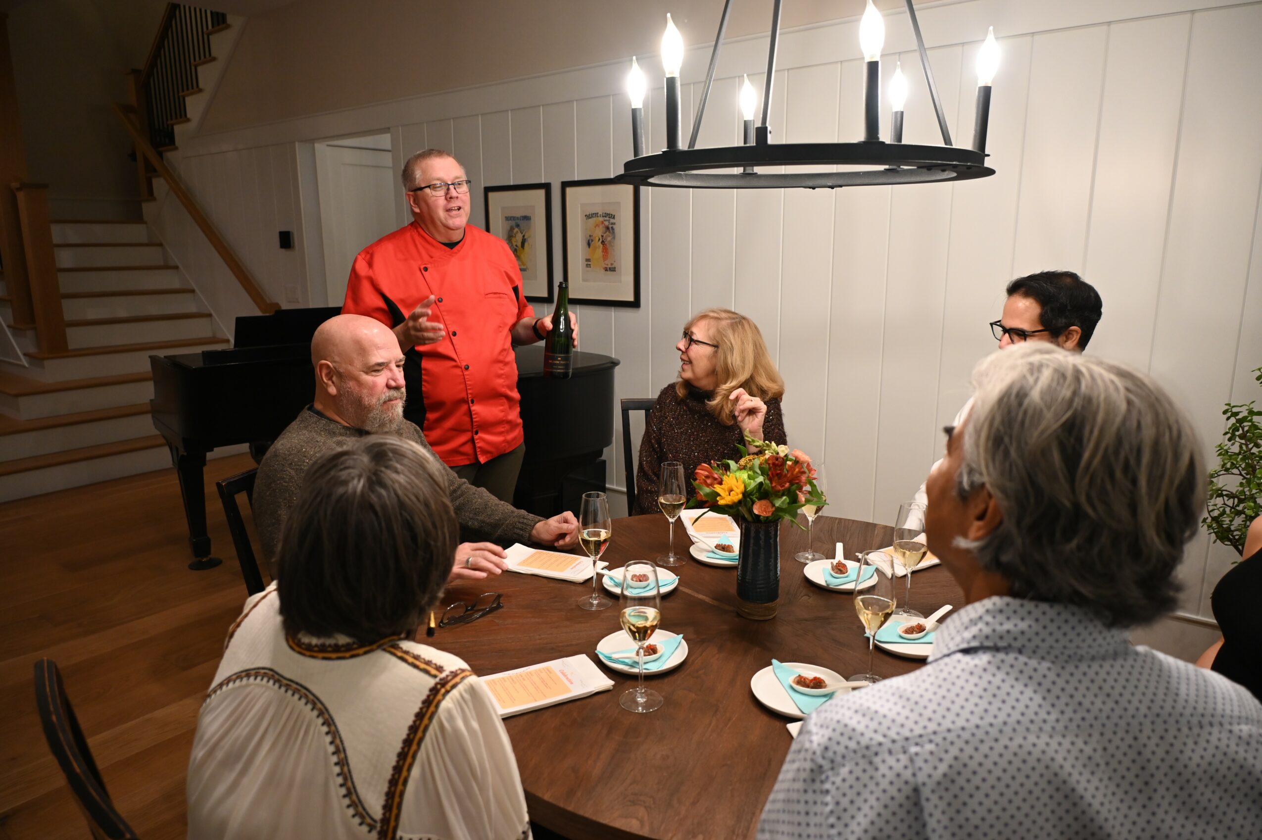 Group of six people gathered around a dining table with tea cups and a flower arrangement, in a well-lit room.