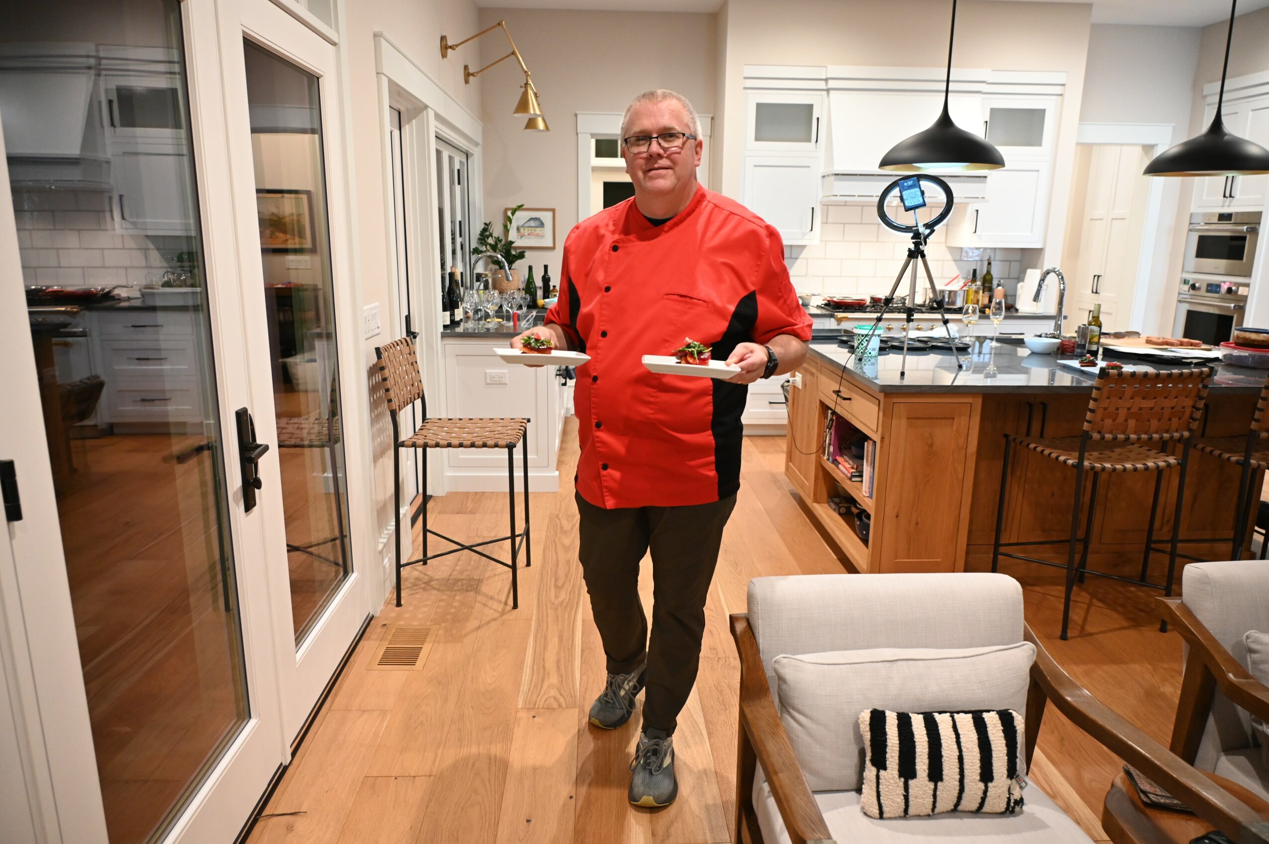 Man in red chef's jacket holding a tray in a modern kitchen with dining area, chairs, and pendant lights.