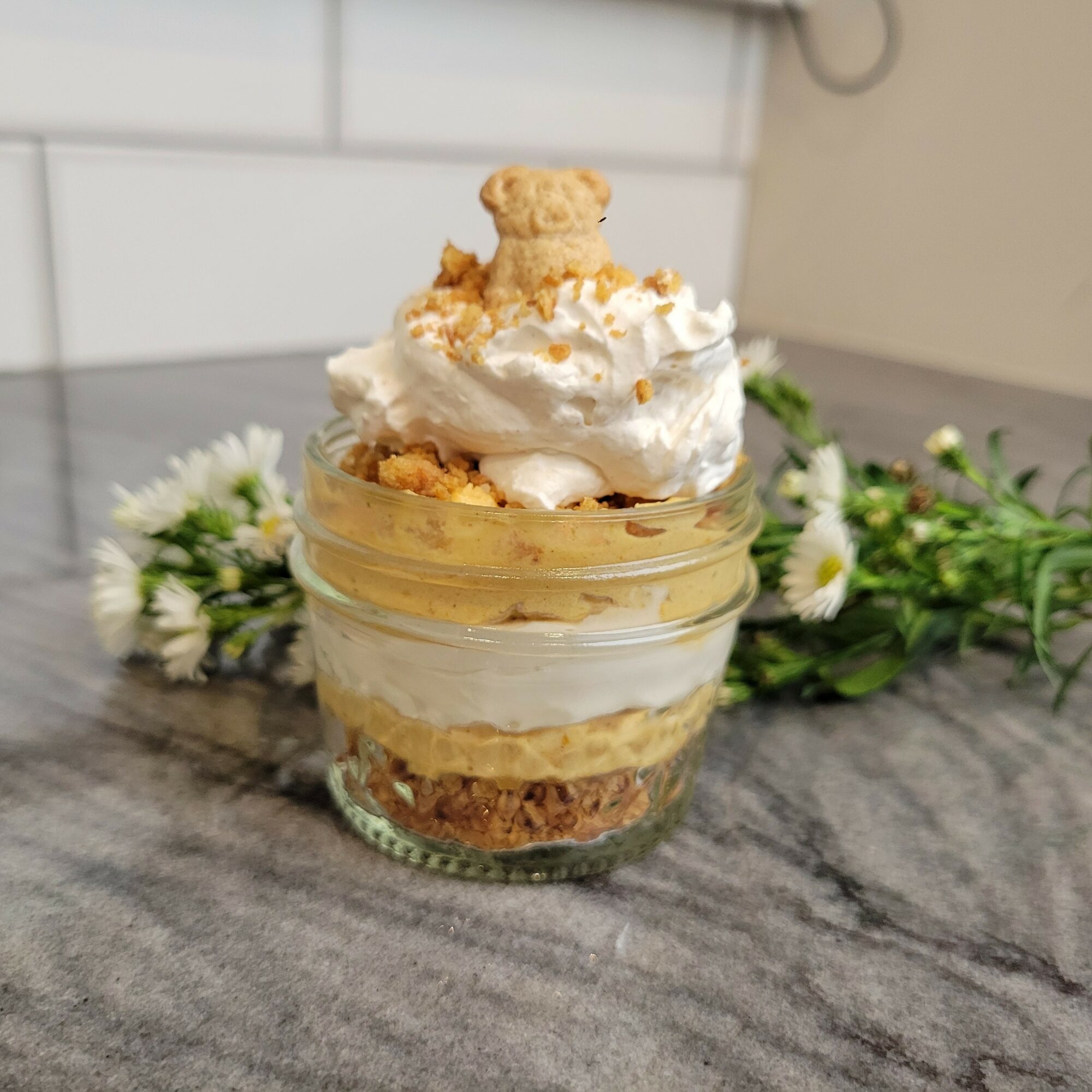 Dessert in a glass jar with whipped cream, cookie crumbs, and a small cookie on top, placed on a marble surface with flowers in the background.
