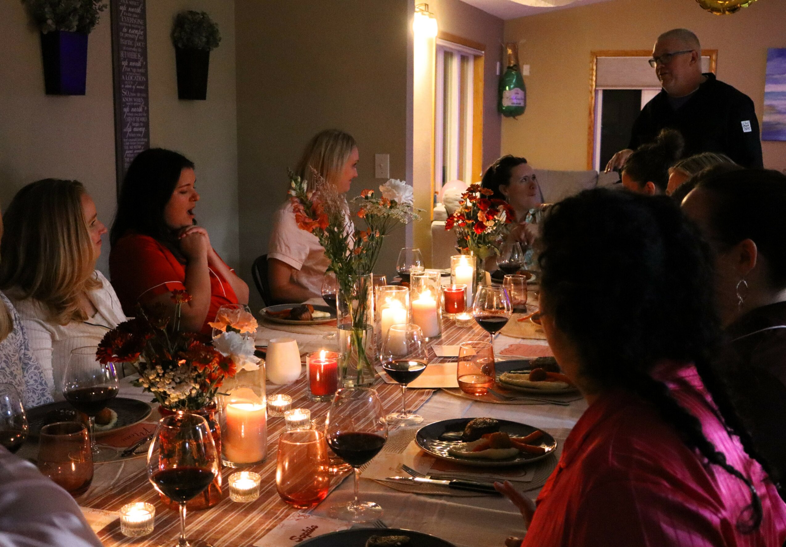 Group of people gathered around a dinner table with candles and flowers, listening to a person standing and speaking.