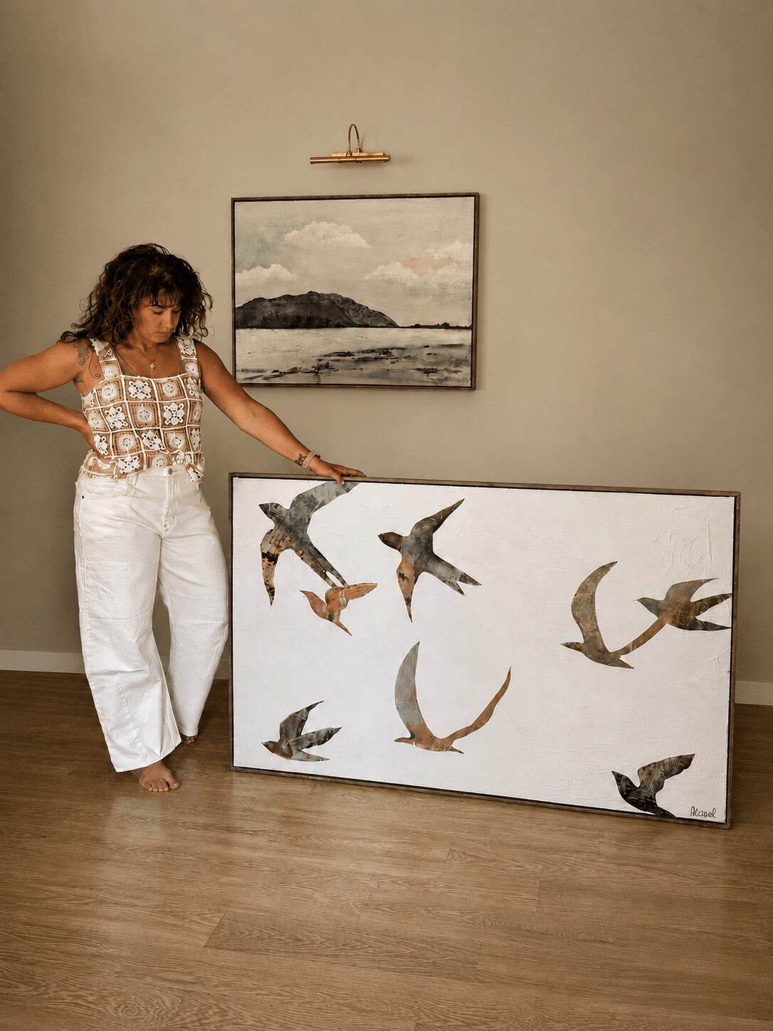 Woman with curly hair in white pants and patterned top stands next to a large painting of flying birds, leaning against a wall.