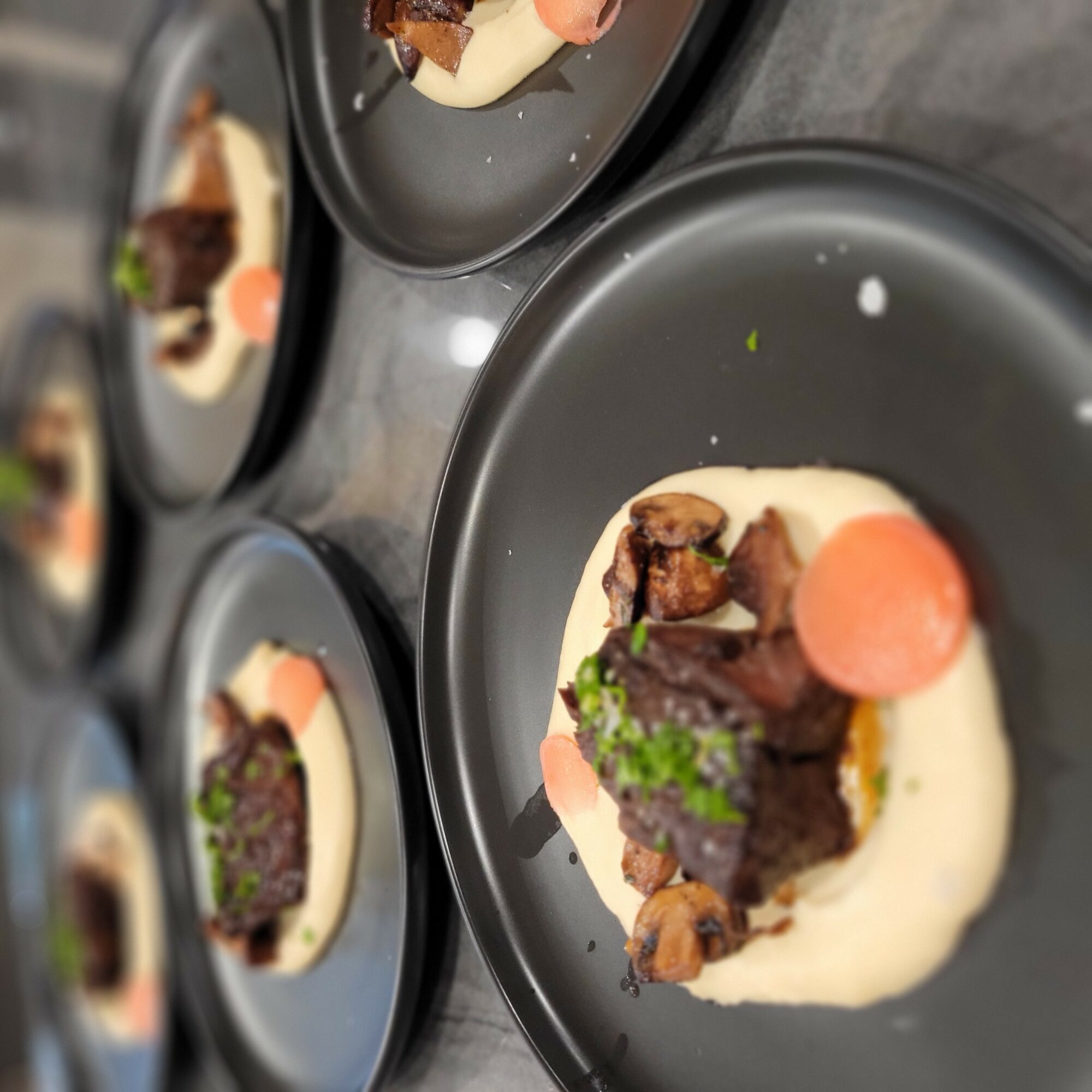 Plate with a piece of cooked meat, a pink sphere, green garnish, and creamy sauce on a black plate.