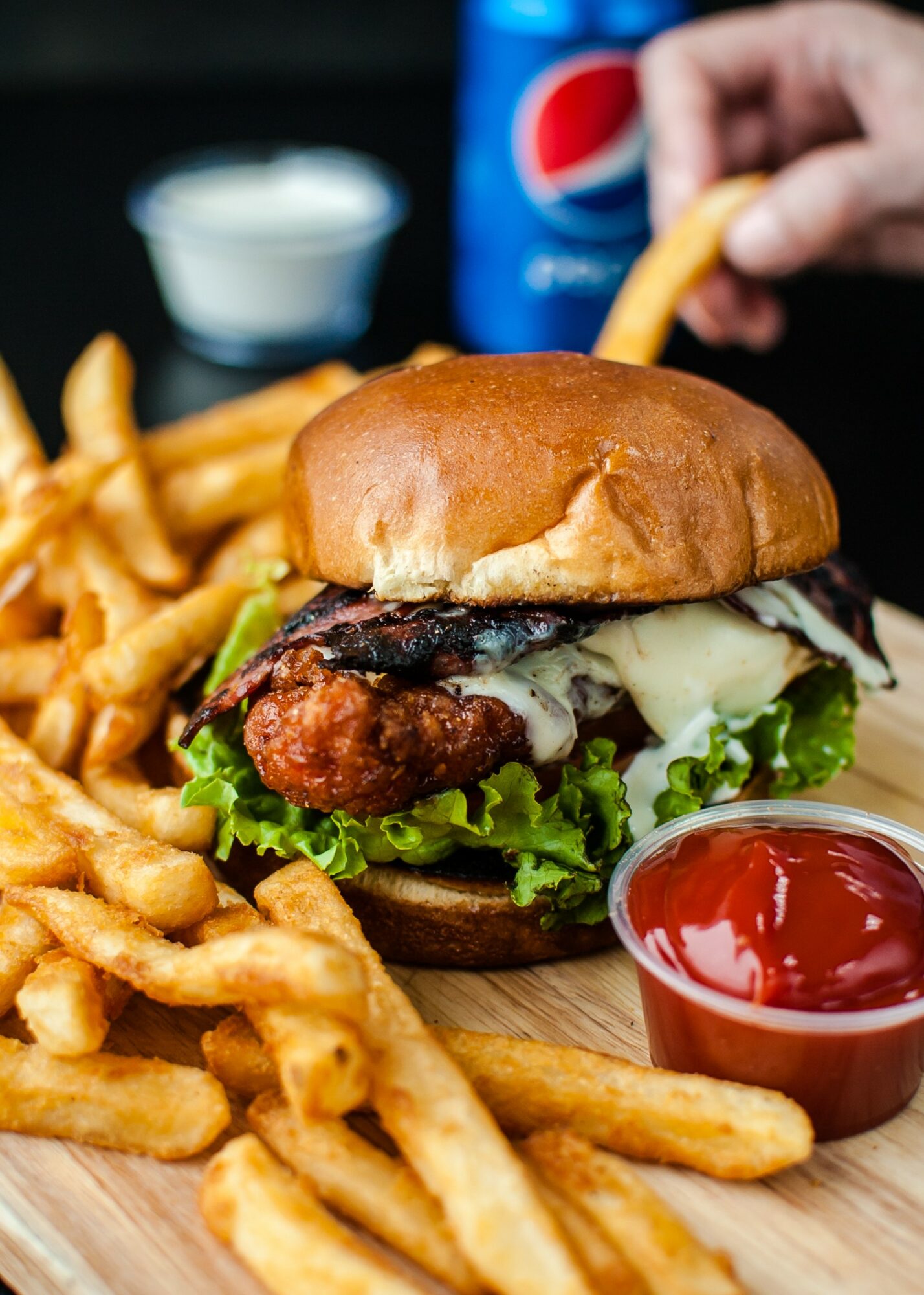 Burger with lettuce, fried chicken, cheese, and bun, served with fries and ketchup on a wooden plate.