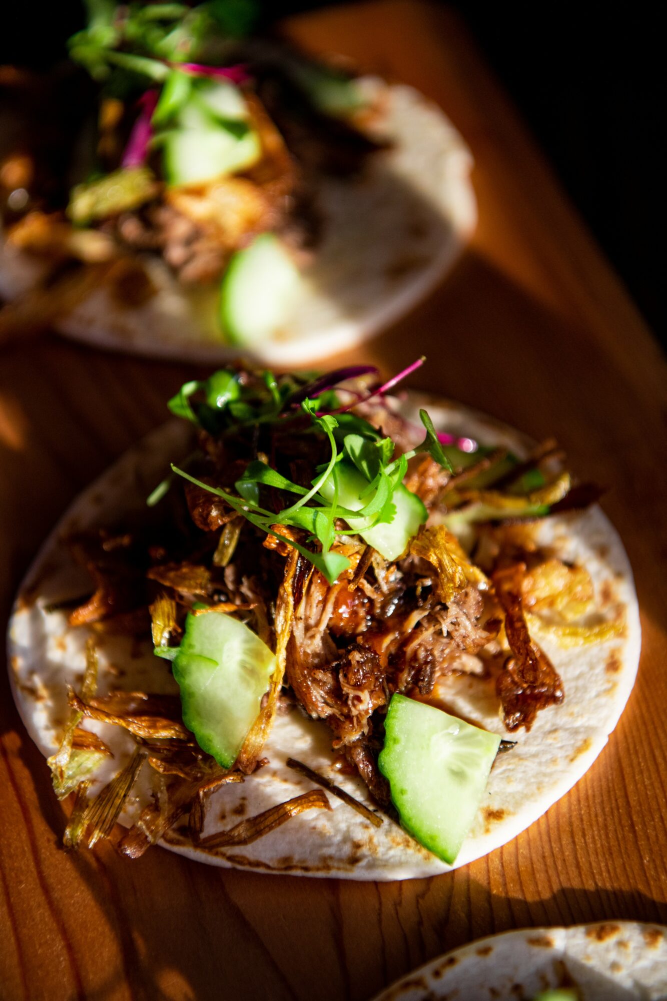 Close-up of a taco with shredded meat, cucumber slices, and microgreens on a wooden surface.