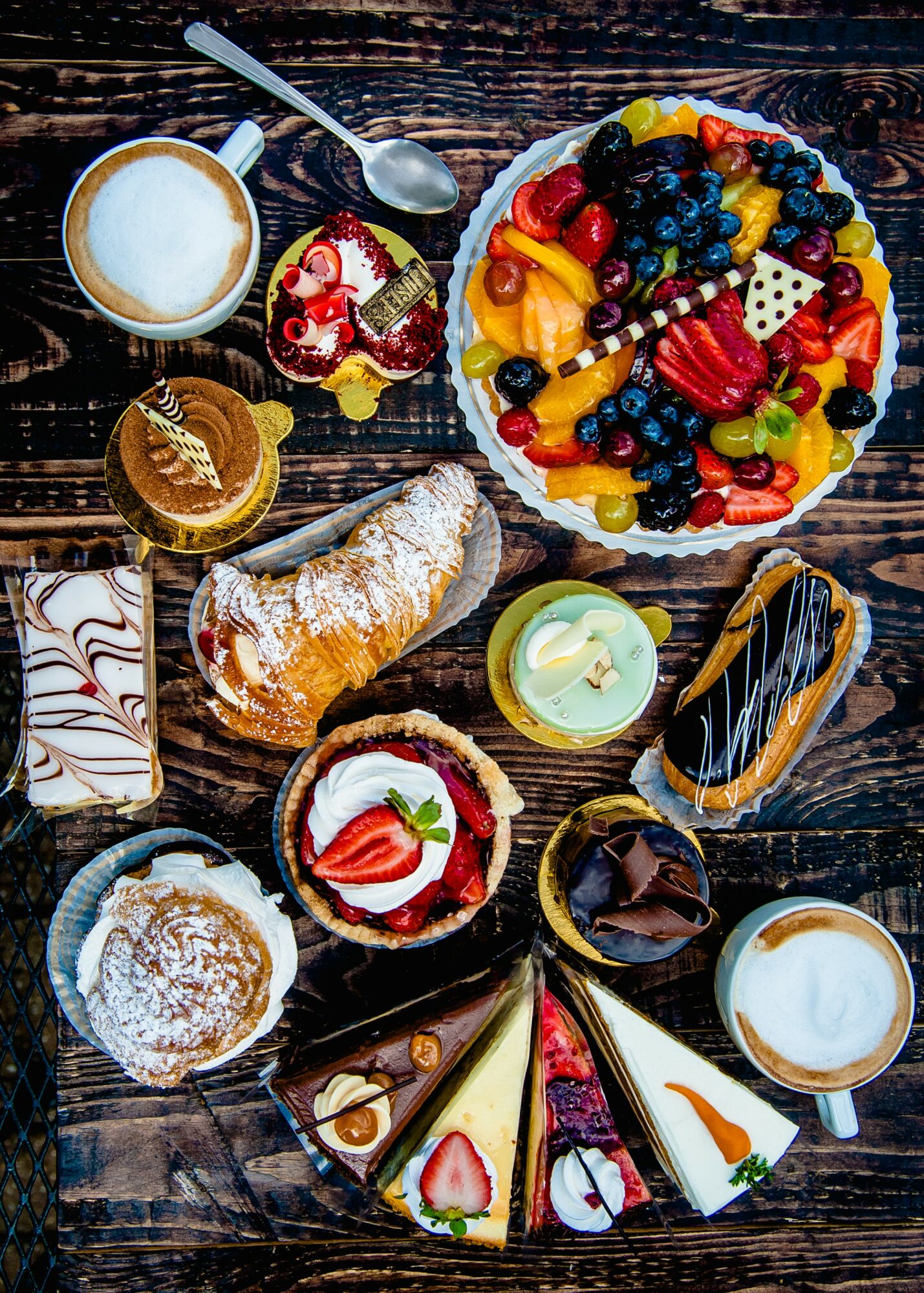 Assorted desserts and a fruit tart on a dark wooden table, including cakes, pastries, and bowls of cream and fruit.