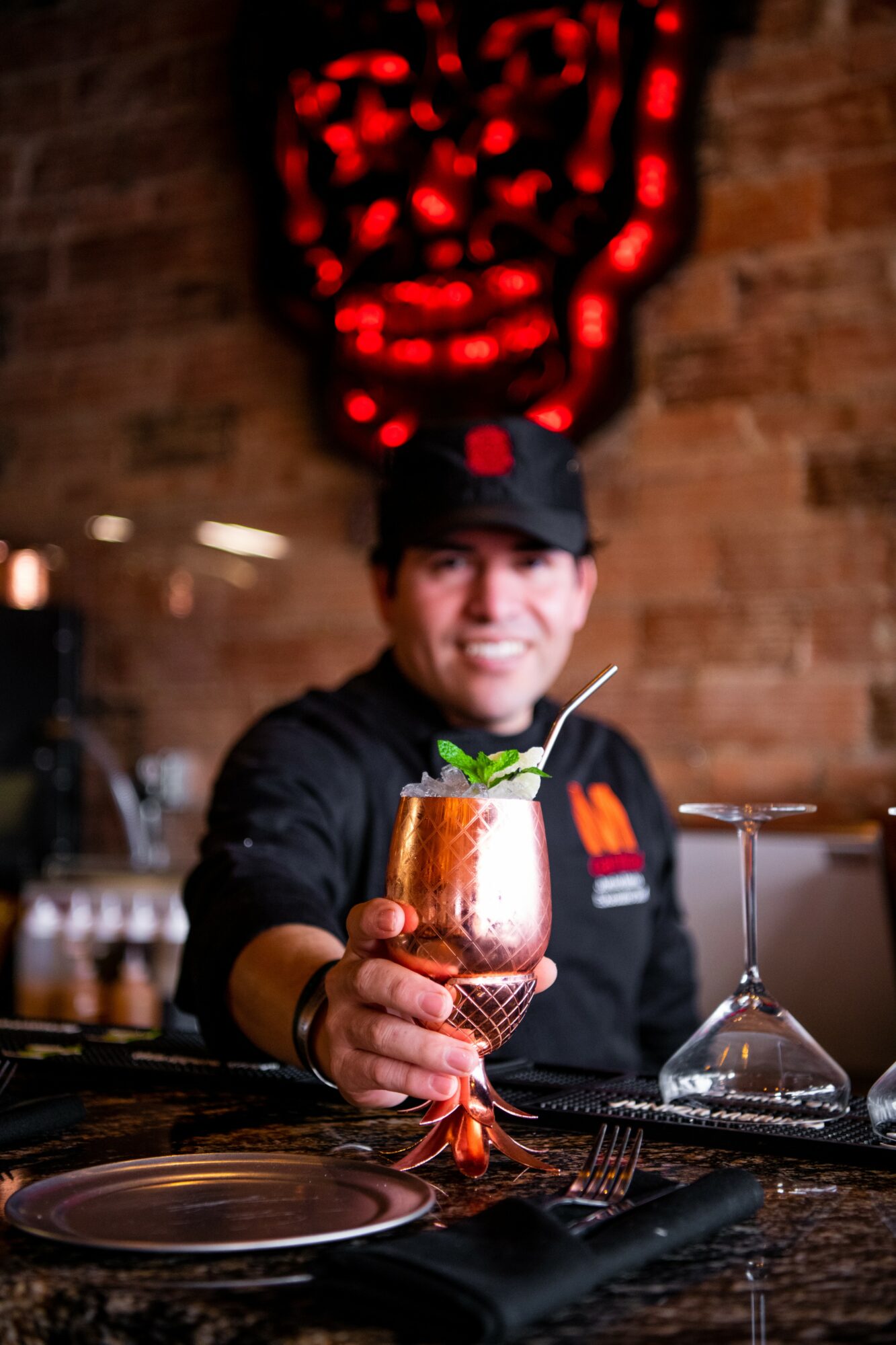 Person in black uniform holding a copper cocktail glass with mint and lime, smiling, brick wall background.