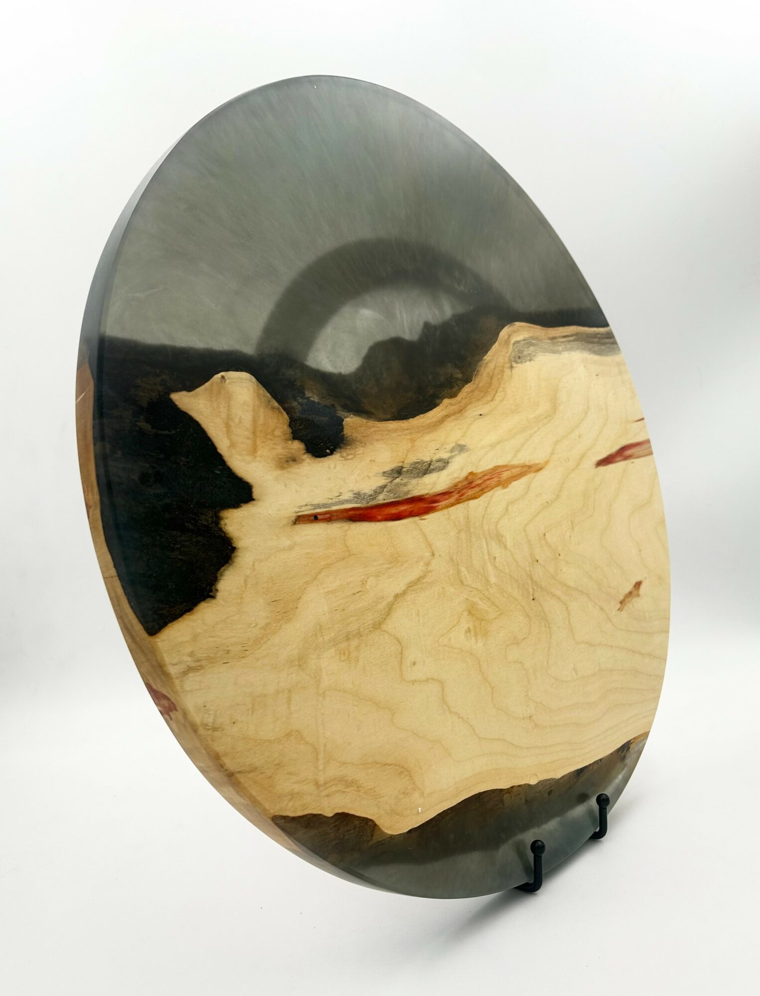 Round wooden art piece with black, beige, and reddish-brown patterns, displayed on a stand against a white background.