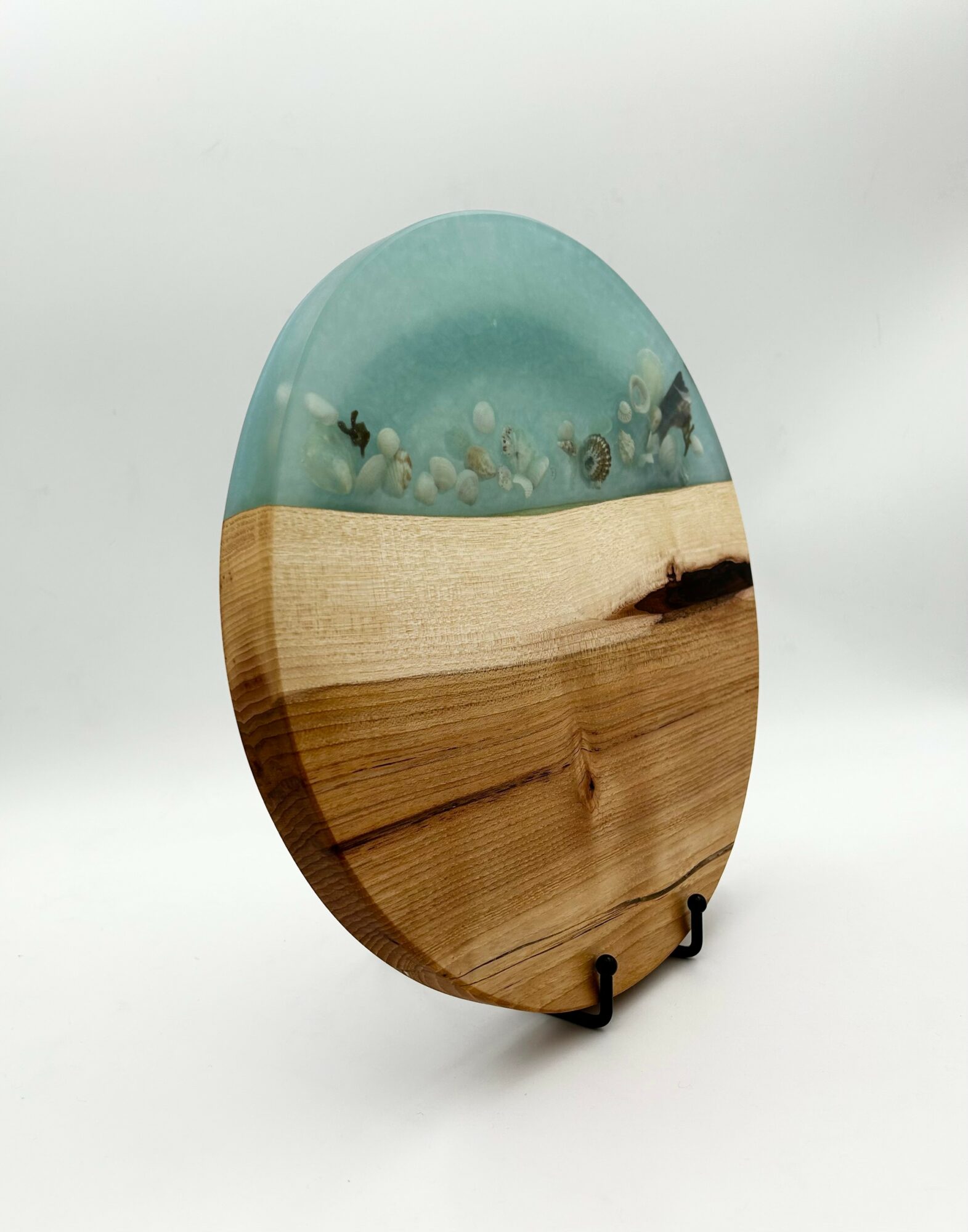 Decorative oval wooden and resin art piece with a beach scene, sand, and water, displayed on a stand.