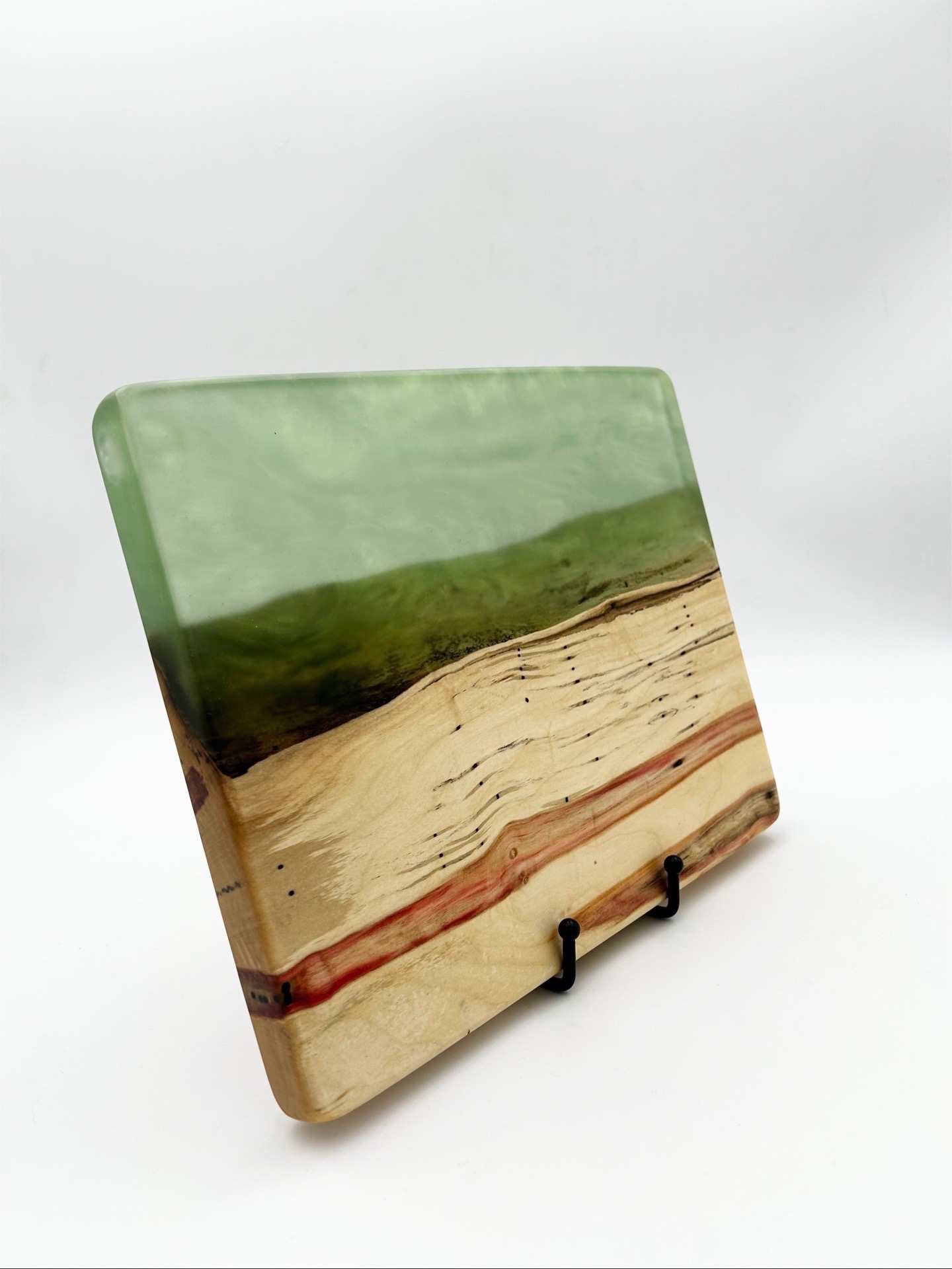 Square decorative stone with layered colors, green top, beige middle, reddish bottom, on a black stand against a white background.