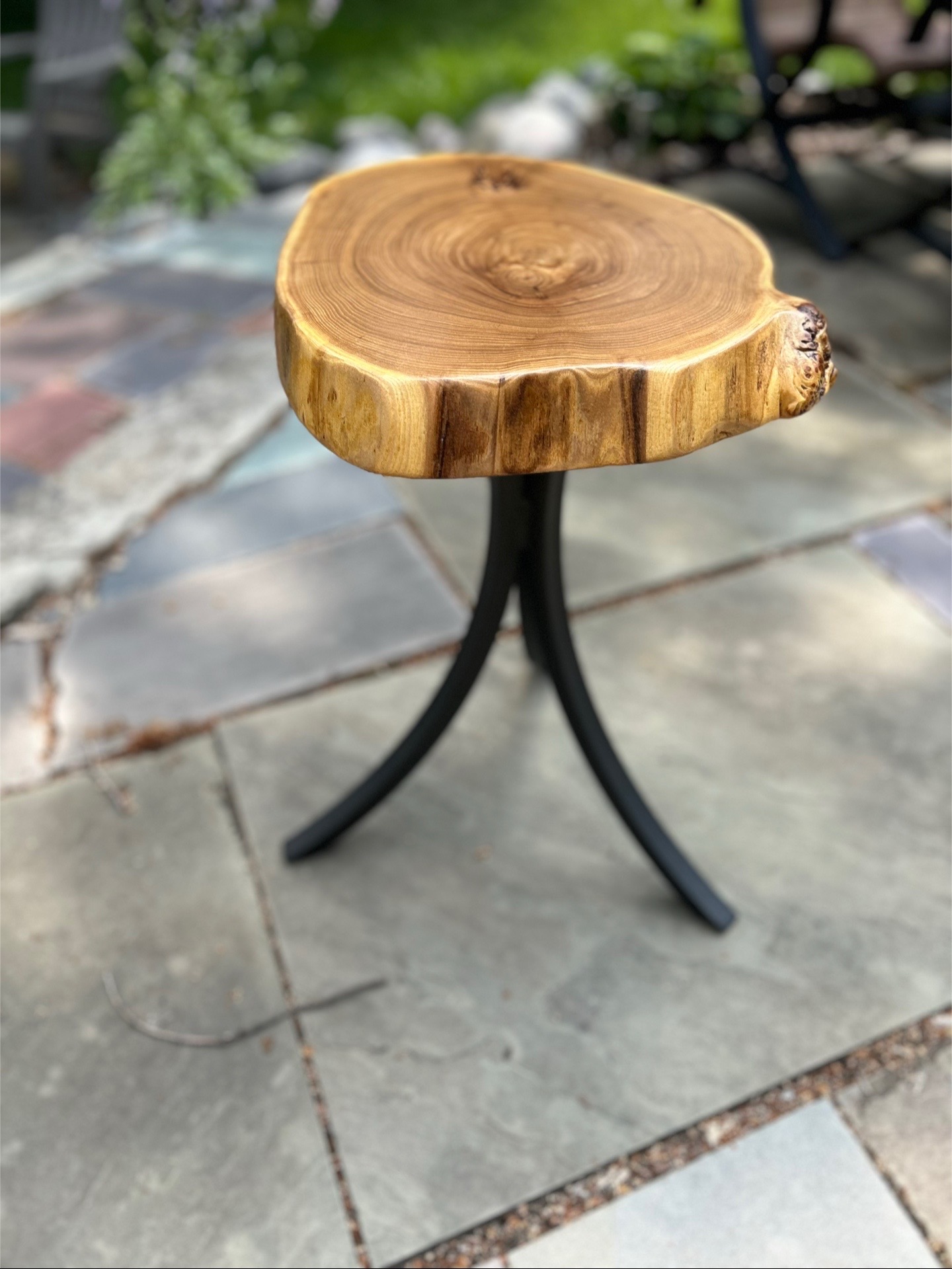Small table with a natural wood top and three black curved legs, outdoors on stone pavement.