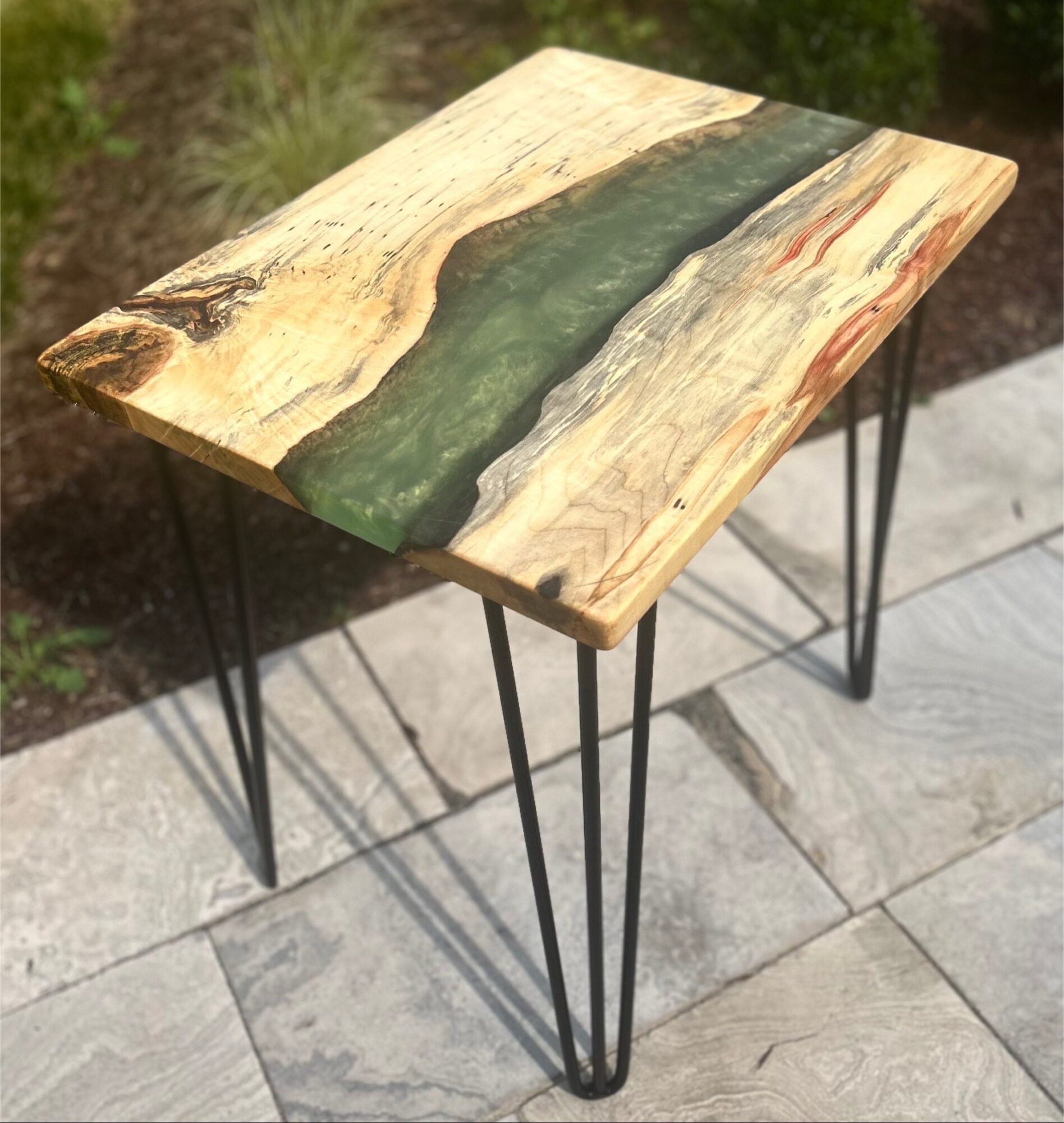 Wooden table with a green resin inlay, standing on black metal hairpin legs, outdoors on stone pavement.